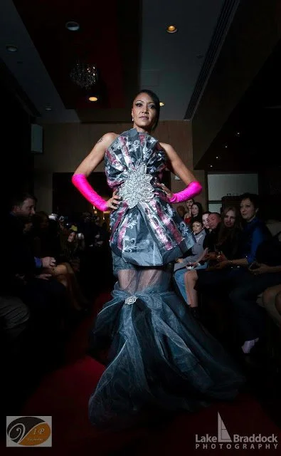 Model walking on runway in an elaborate dress with layered fabric and a long tulle train, wearing bright pink gloves, at a fashion show.