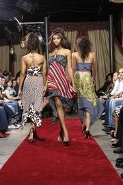 Three models walking down a runway during a fashion show, wearing colorful dresses, with an audience seated on either side.