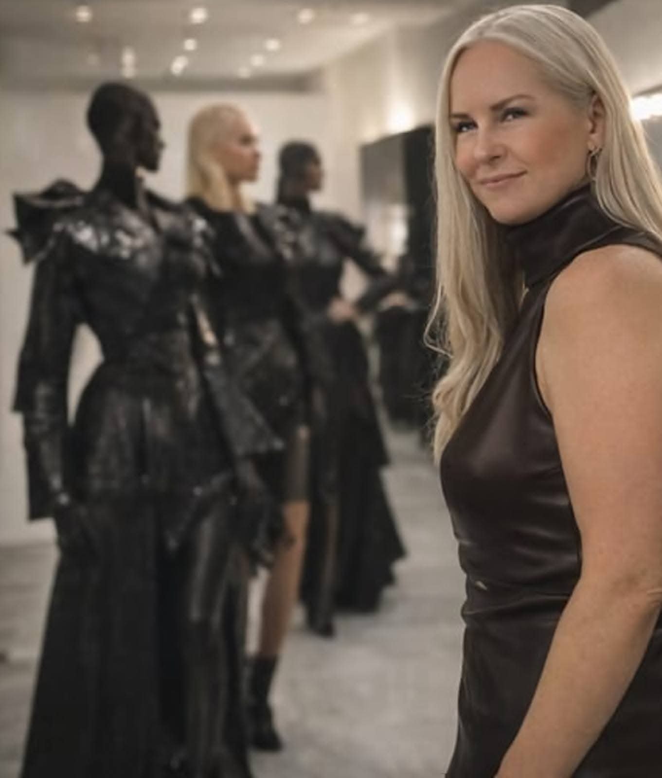 A blonde woman in a shiny black dress smiling, with four women in black futuristic outfits with armor-like shoulders blurred in the background.