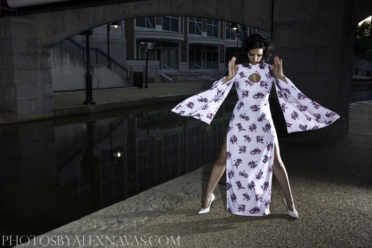 Woman wearing a long white dress with purple Chinese characters, high heels, standing on a sidewalk near a canal under a bridge in an urban area.
