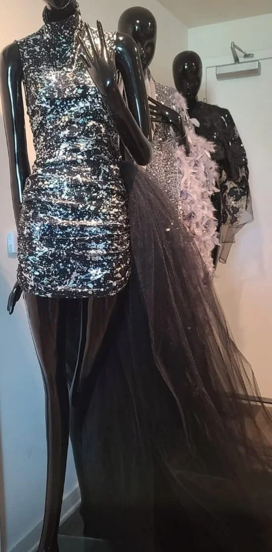 Display of mannequins dressed in glamorous, high-fashion outfits, including a sparkly silver dress and a black tulle gown with feathered details.