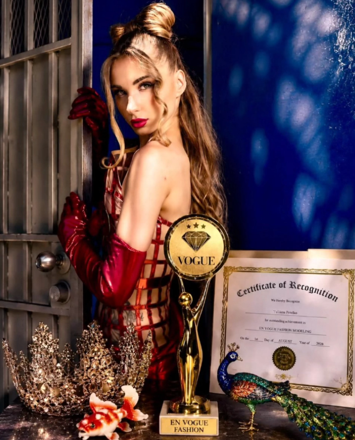 A woman dressed in a red, cage-style dress with long red gloves, posing behind a table with awards and decorations, including a gold trophy labeled 'EN VOGUE FASHION', a diamond plaque with 'VOGUE', a certificate, a peacock figurine, rhinestone crowns, and a goldfish.