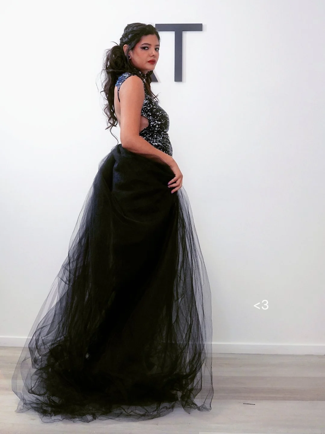 Young woman in a black sequined top and a voluminous black tulle skirt standing against a plain white wall with a 'T' letter on the wall and a small heart symbol near the floor.