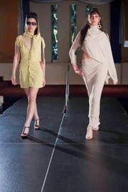 Two female models walking on a runway during a fashion show, wearing light-colored outfits and sunglasses.