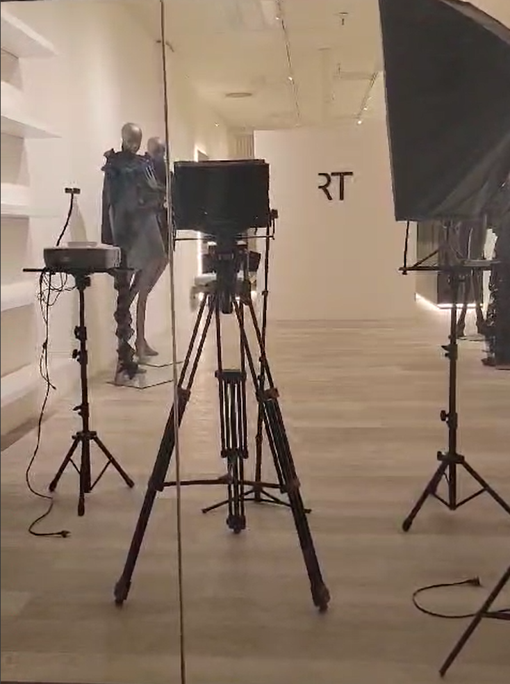 View of a studio or gallery space with two mannequins dressed in dark clothing, set against a mirror that reflects the room, with the letters 'RT' on the wall in the background. Several camera and lighting setups are positioned in the foreground.