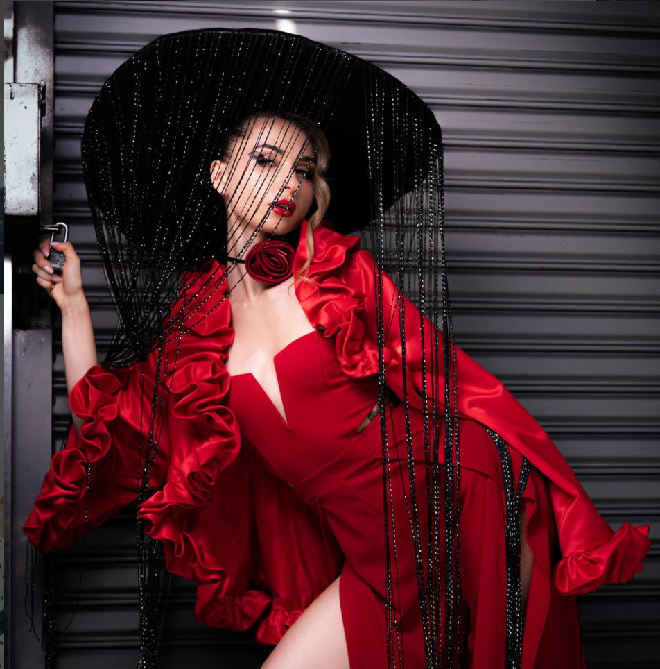 Woman in a red dress with ruffled details and a large black hat with beaded fringe, standing against a shuttered background.