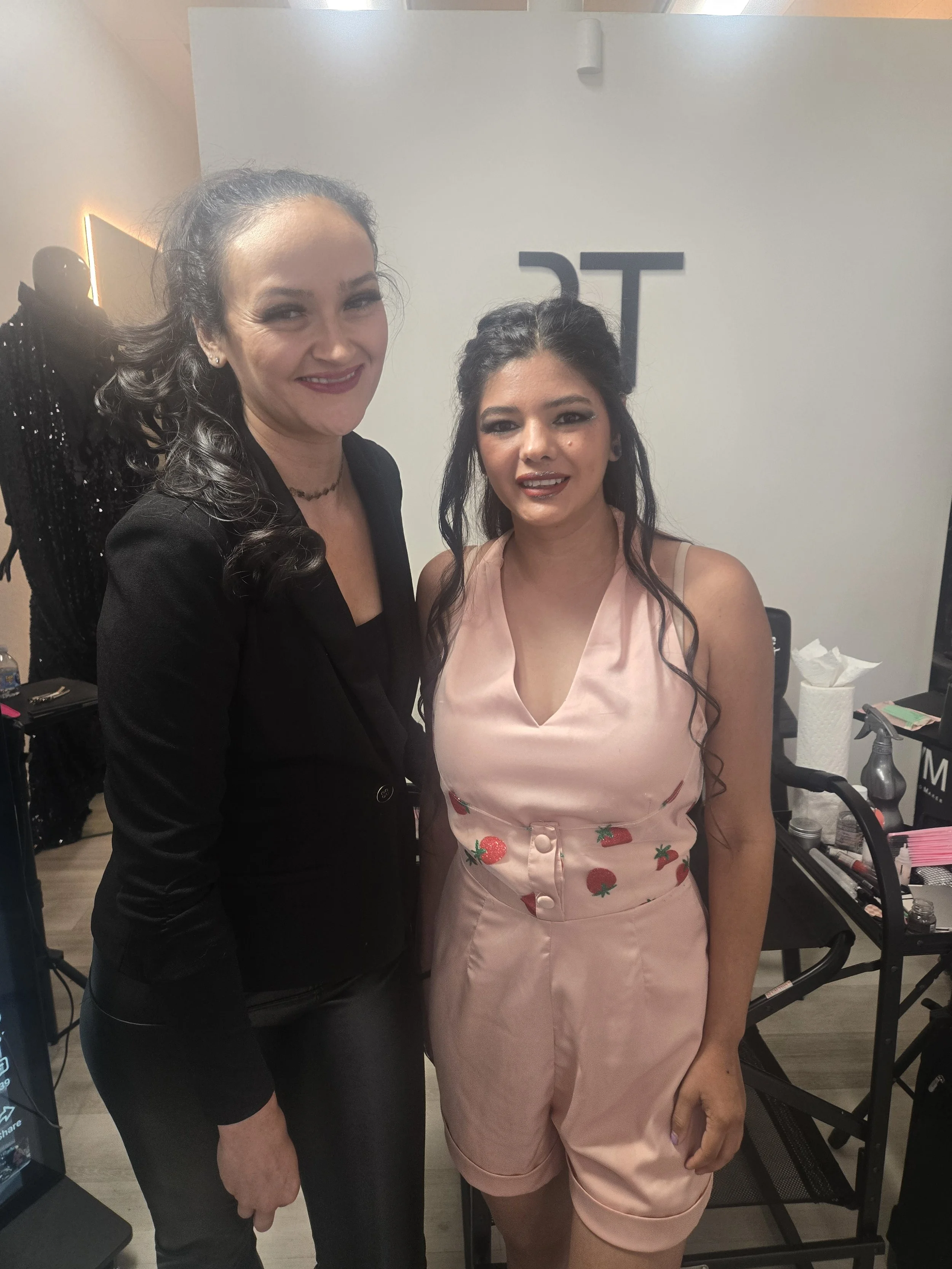 Two women posing together at an indoor event, one with dark curly hair and makeup wearing a pink dress with strawberry embroidery, the other with dark hair in a bun, wearing black clothing and a choker, standing in front of a white wall with the part