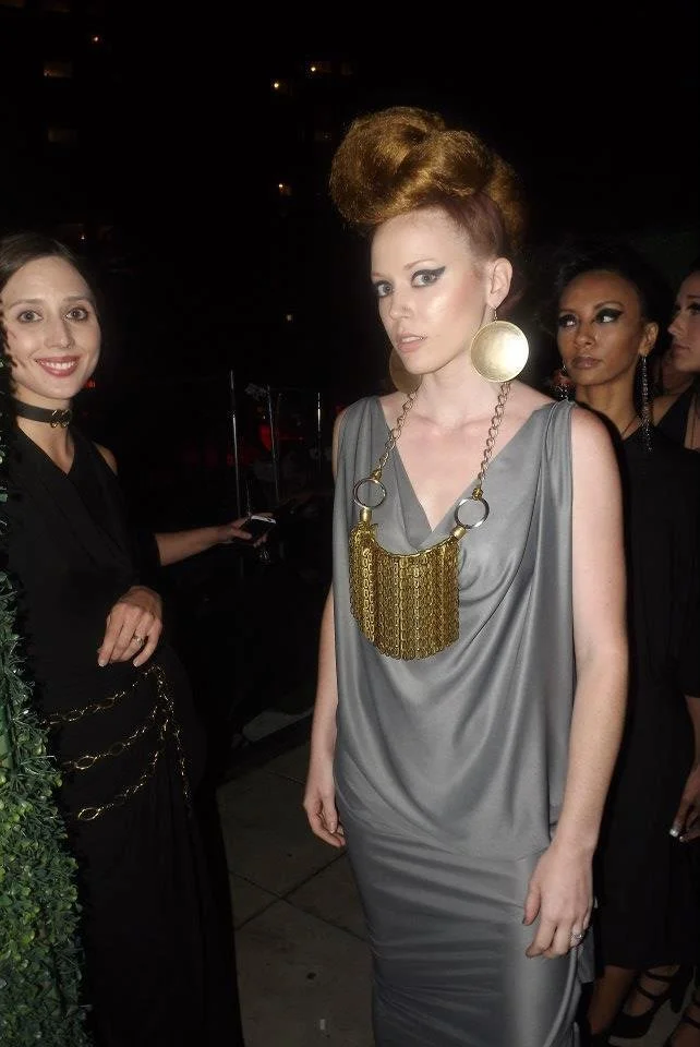 Group of women at a social event, one woman in the foreground wearing a silver satin dress with bold jewelry, others in black dresses in the background.