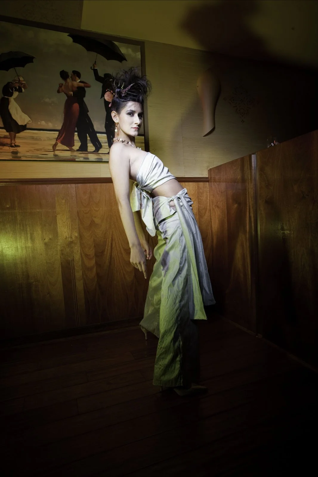 A woman wearing a shiny silver dress with a midriff cutout and high heels, standing in a dimly lit room with wood paneling, and a large framed painting of people dancing and holding umbrellas hanging on the wall behind her.