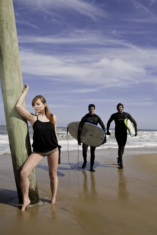 Woman in black top and shorts leaning against a wooden pole on the beach, two men in wetsuits carrying surfboards walking toward the ocean.