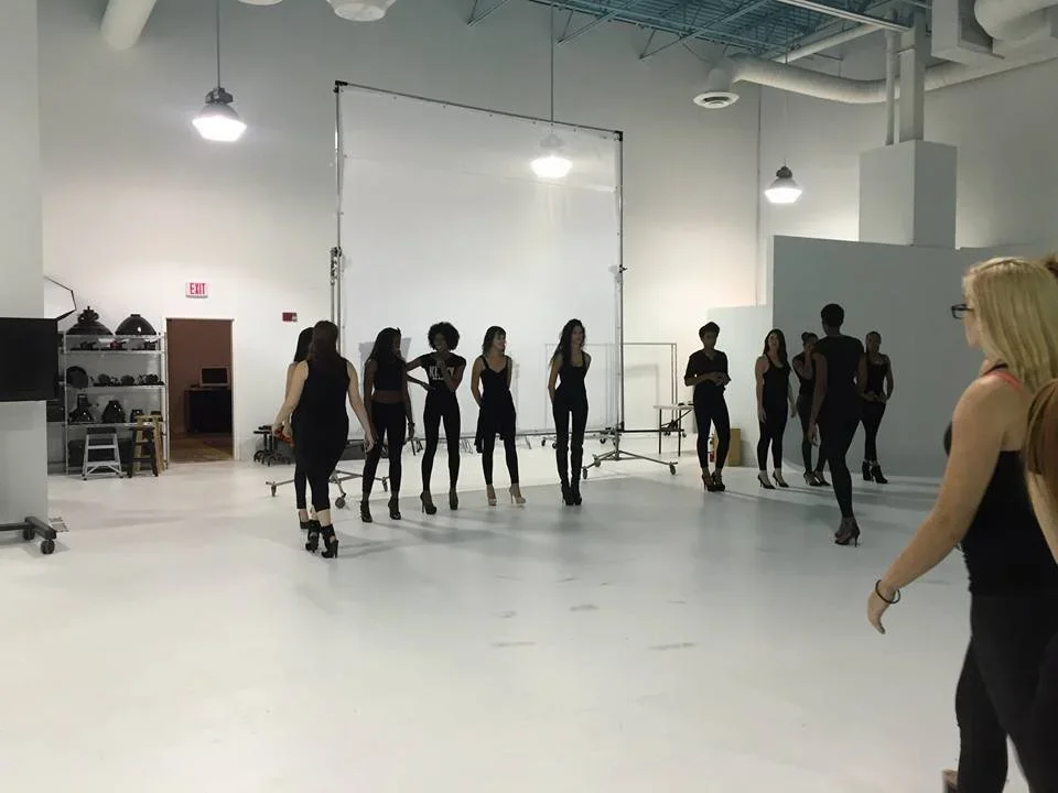 Group of women in black dancing or practicing in a spacious dance studio with white walls and high ceilings.