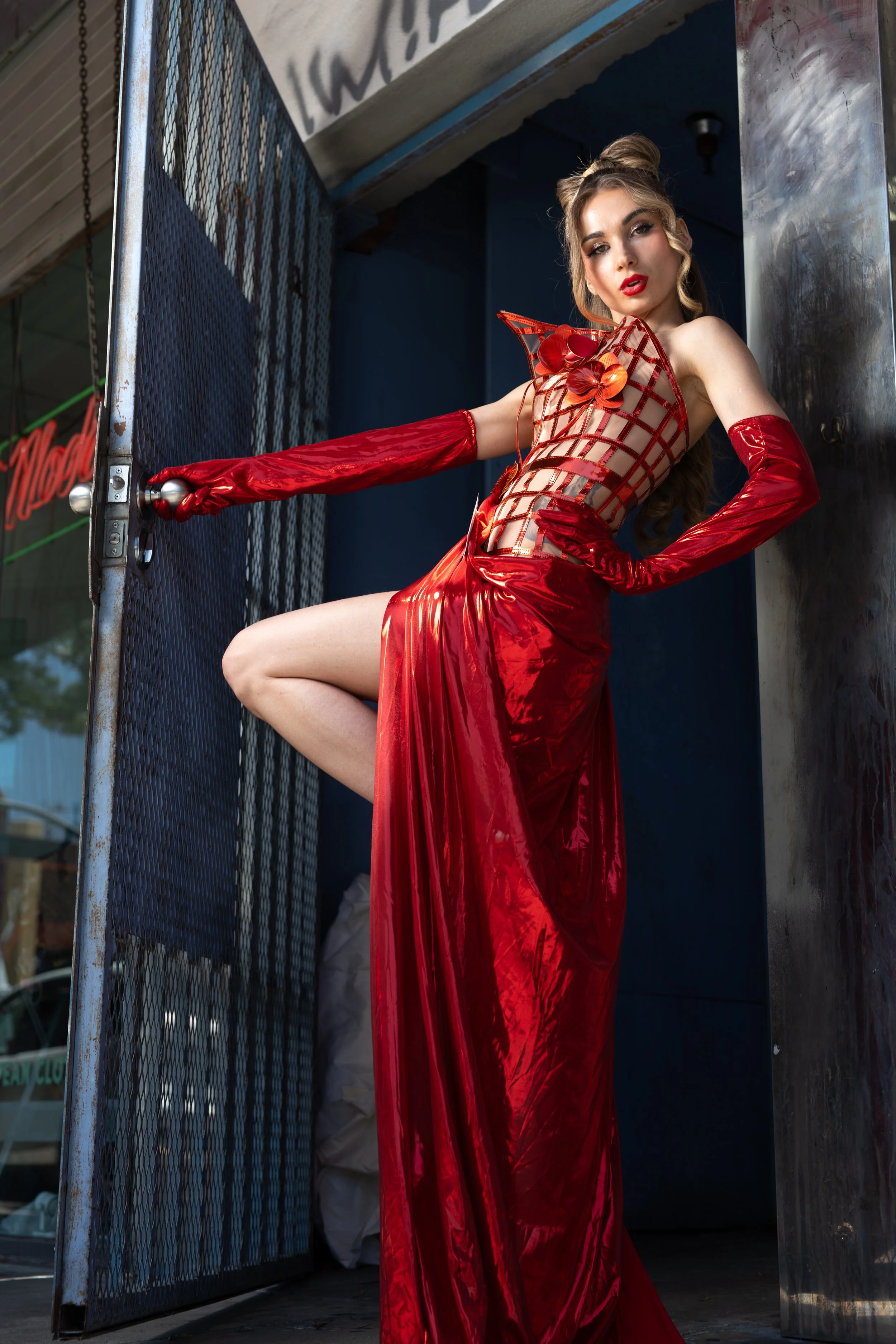 Woman in a red, sheer dress with floral embellishments, standing next to a metal gate, looking at the camera with a confident expression.