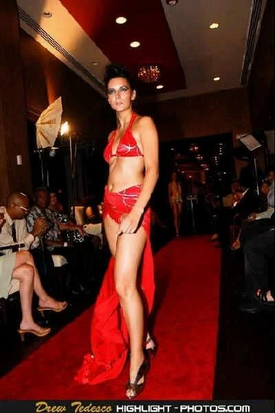 A woman walking down a runway in a red outfit surrounded by seated spectators in an indoor venue.