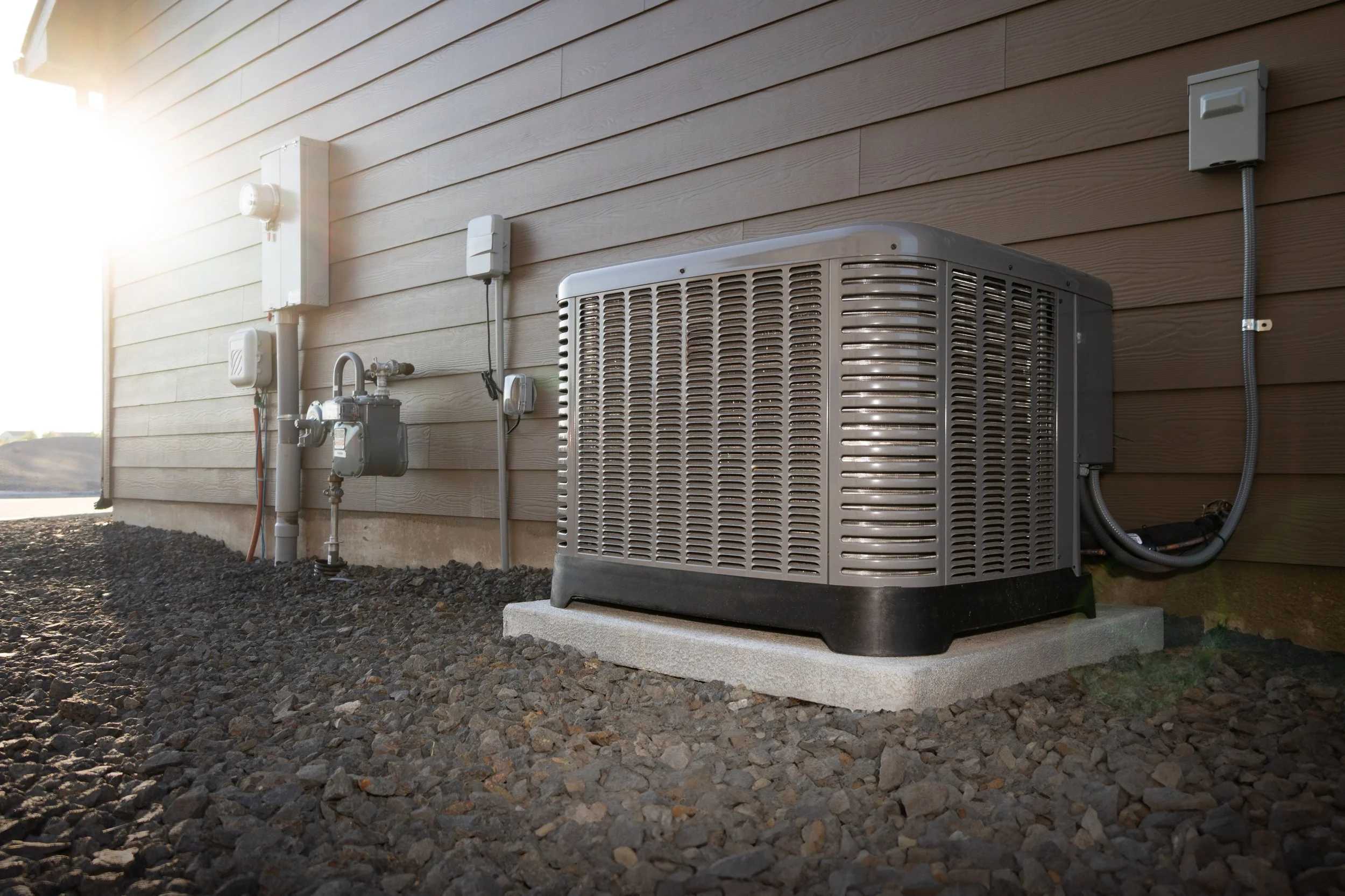 An outdoor view of an air conditioning unit installed next to the beige siding of a house, with electrical boxes and utility meters on the wall and a gravel ground.