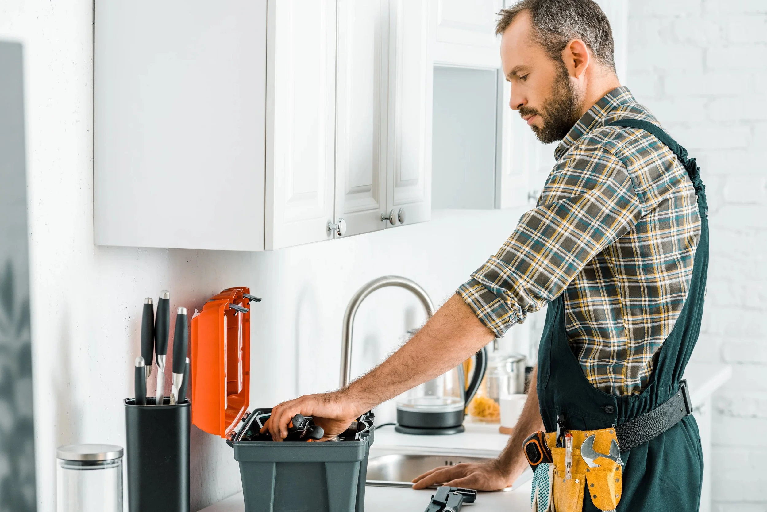 A man wearing a plaid shirt and work apron organizing tools in a kitchen workshop.