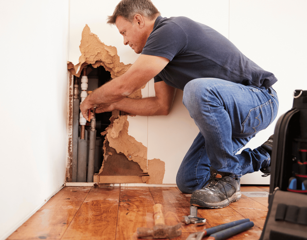 Man repairing plumbing behind a damaged wall with torn drywall and exposed pipes and pipes in the corner.