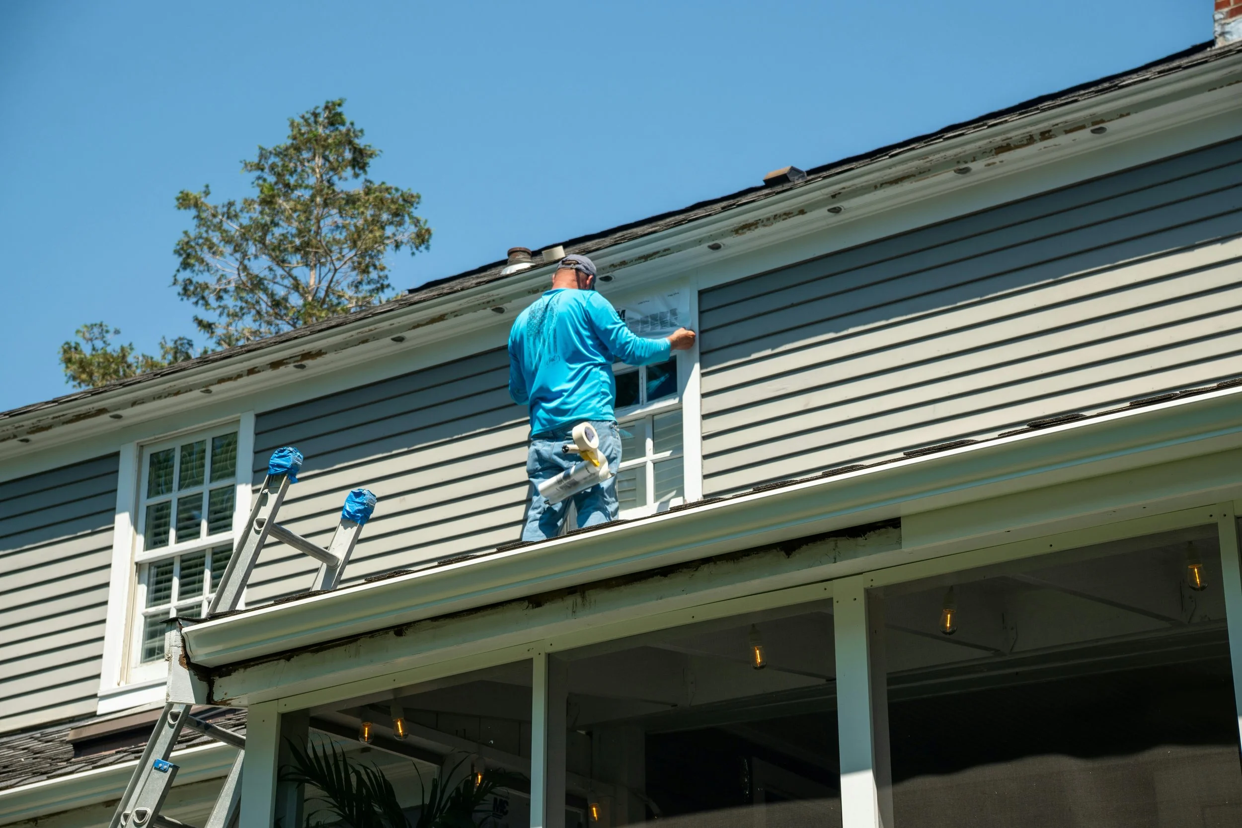 Man working on house siding, standing on ladder, wearing a blue shirt and gray cap, with tools hanging from his jeans, house with gray siding, window, and a tree in the background.