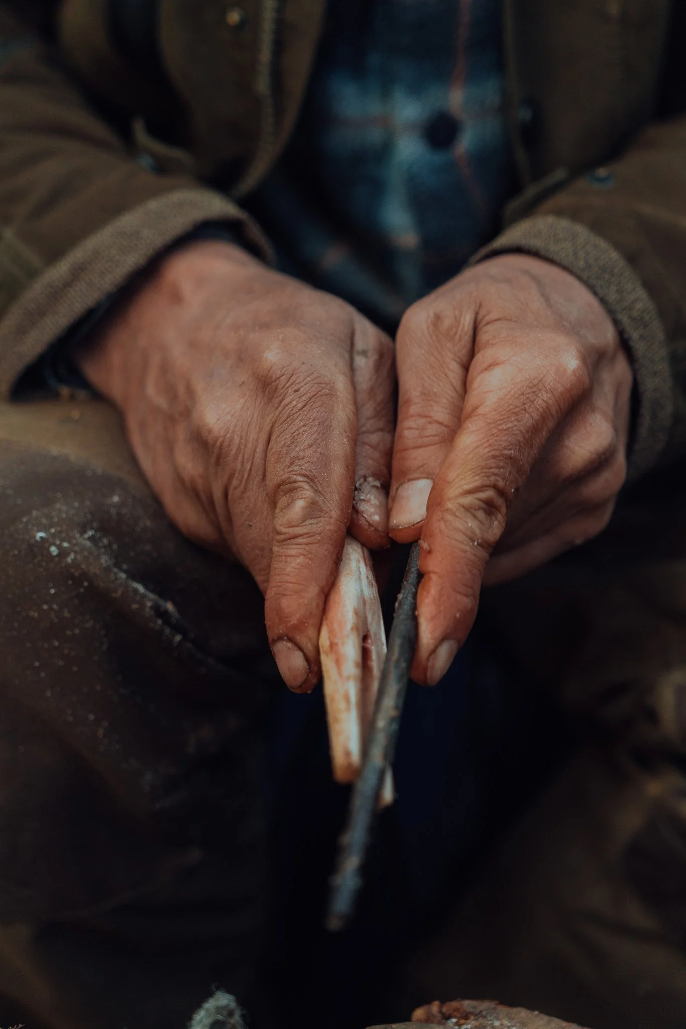 Hands of a person sharpening a knife, wearing a brown jacket and a plaid shirt underneath.