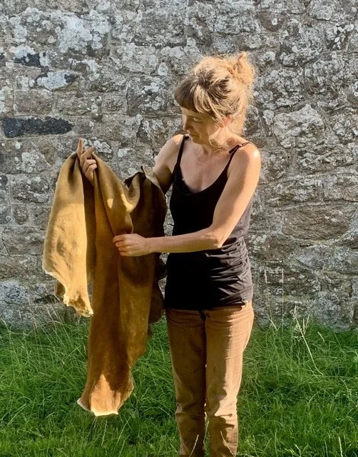 Woman holding a large, tan-colored animal hide or fur in front of a stone wall, standing on green grass.