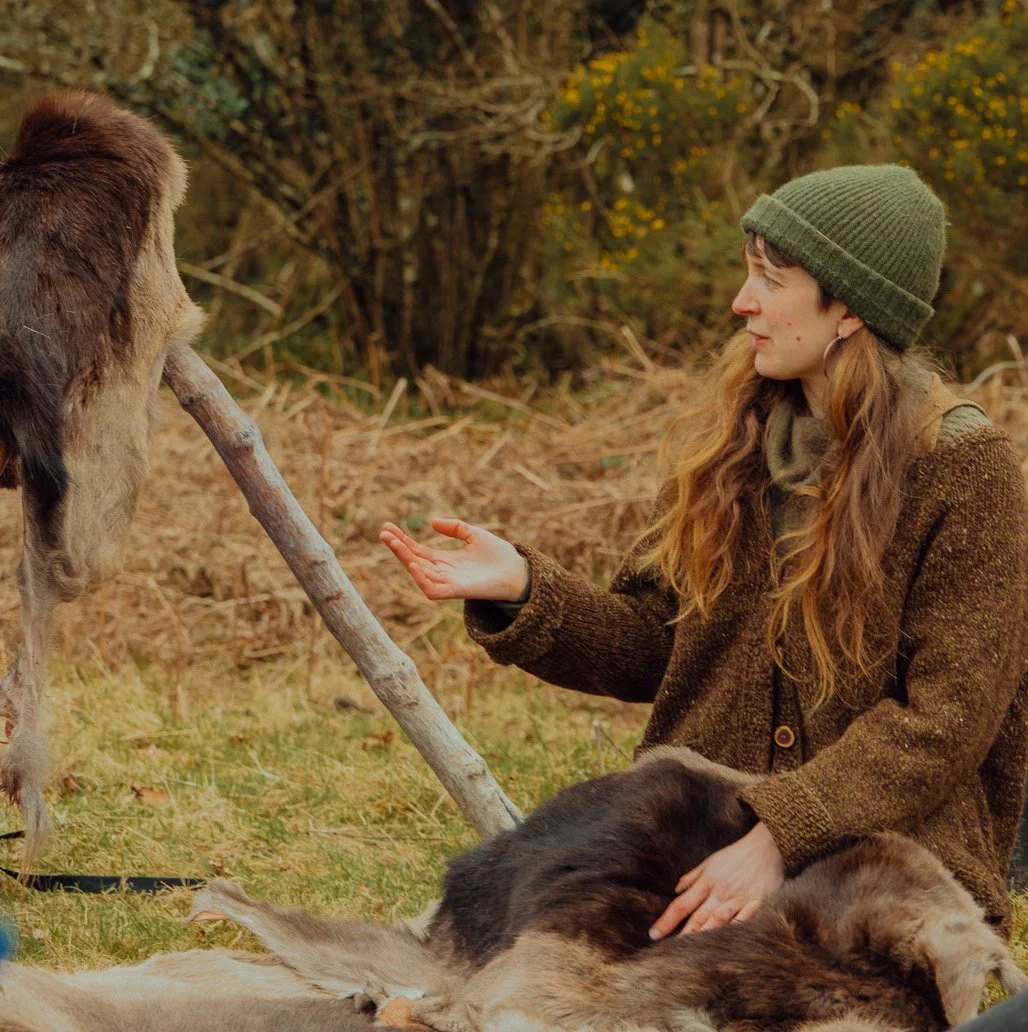 A woman with long red hair wearing a green beanie and brown coat, sitting outdoors, pointing to an animal skin she is holding on a stick, surrounded by trees and autumn foliage.