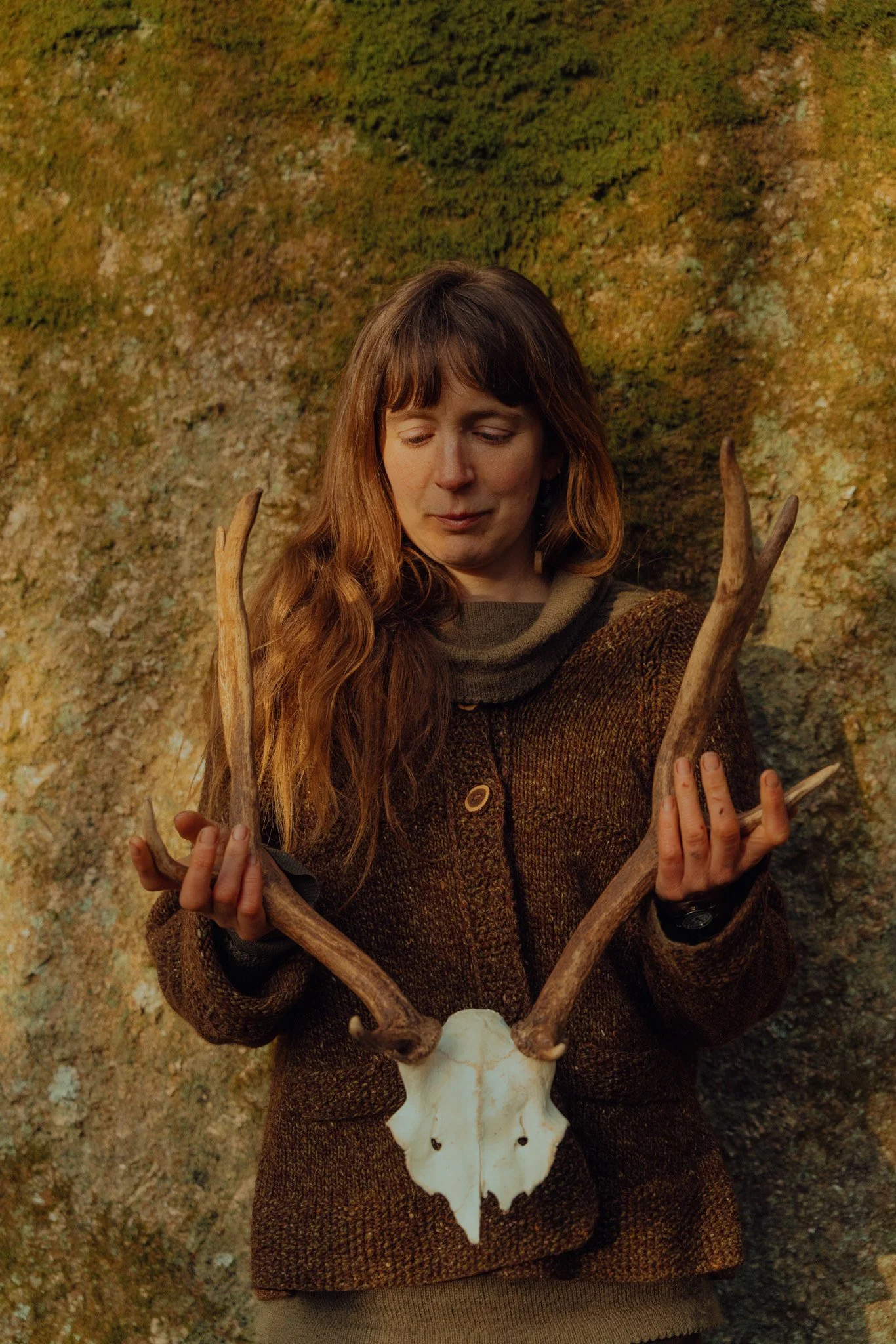 A woman with long reddish-brown hair dressed in a brown sweater and dark watch, standing against a mossy rock background, holding a deer skull with antlers.