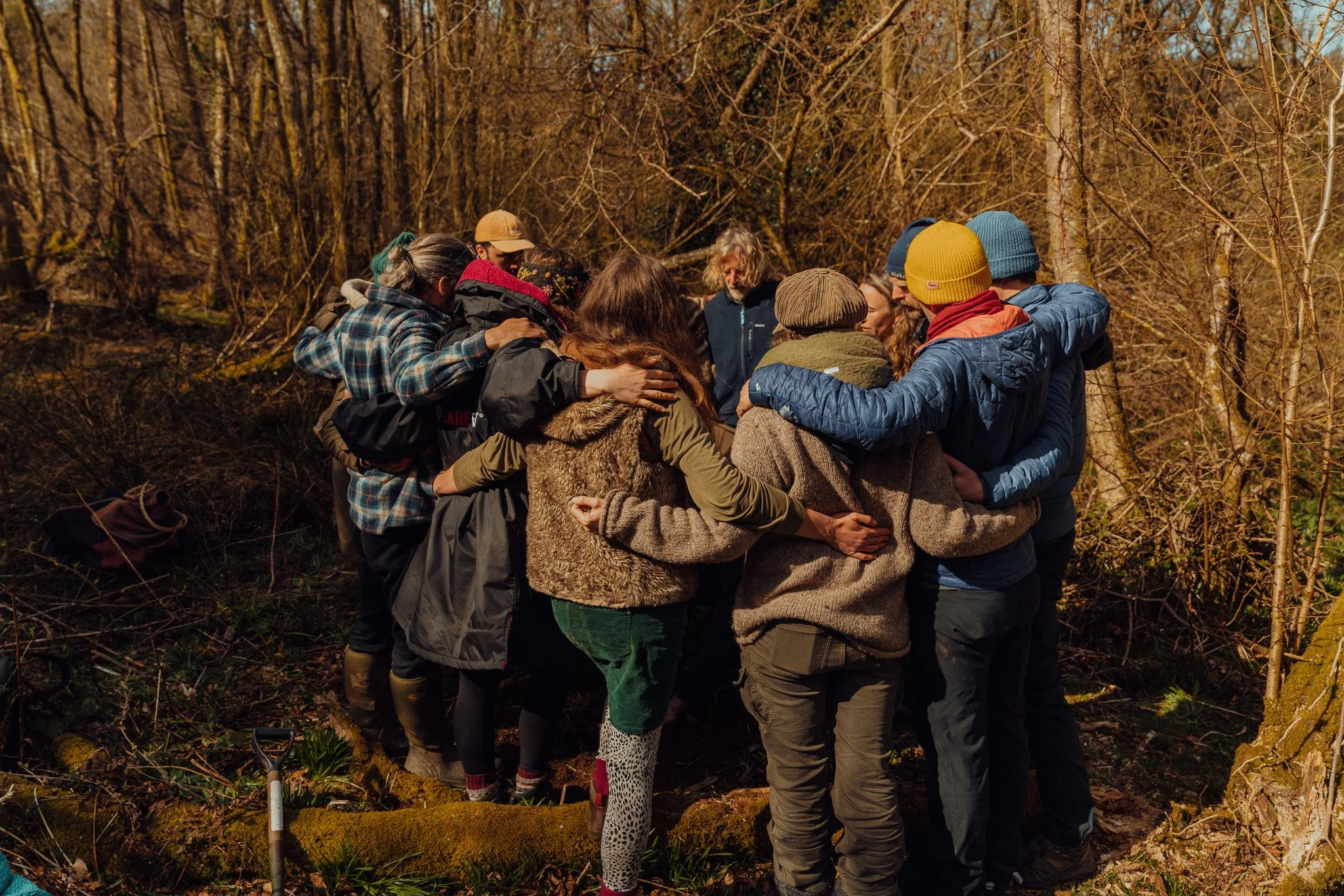 Group of people standing close together outdoors in a circle, with their arms around each other's shoulders, in a wooded area during daytime.