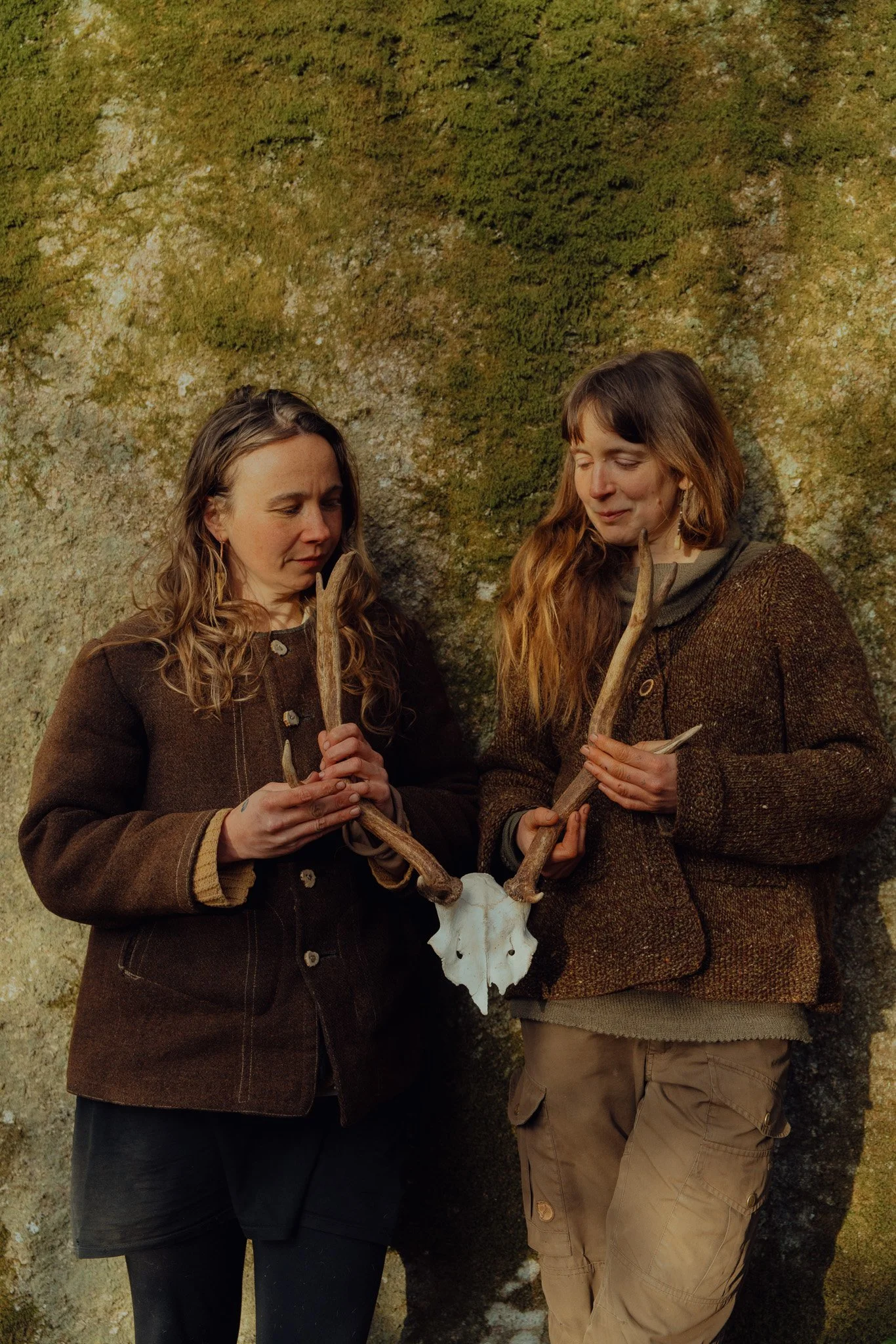 Two women holding antlers and a skull with a mossy rock background.