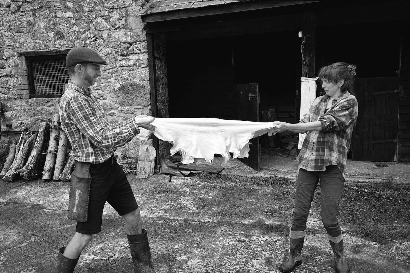 A man and a woman are outdoors, holding a large piece of animal hide or skin between them. They are dressed in plaid shirts and rubber boots, and are stretching a hide between them, with a rustic barn or shed in the background.