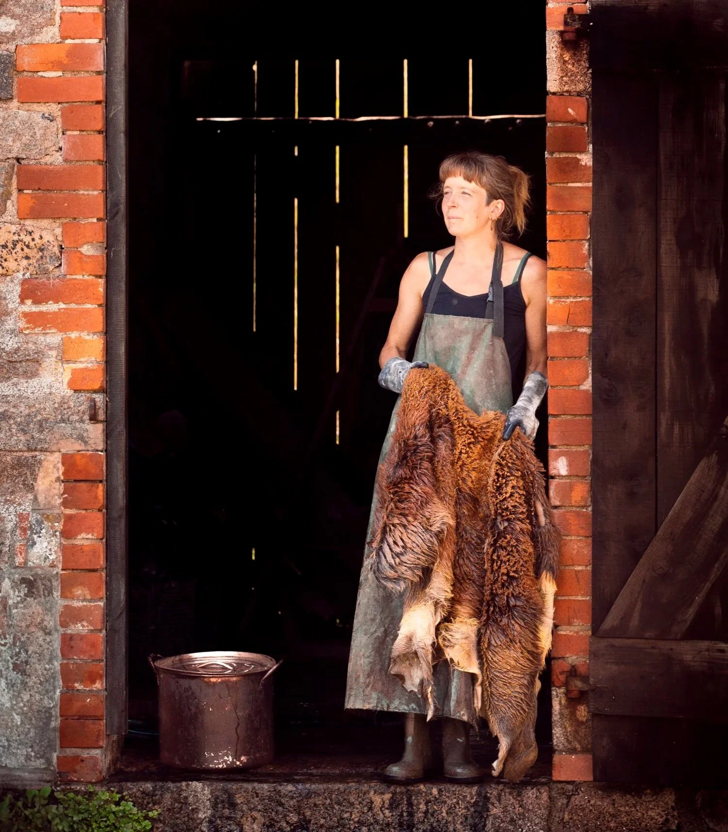 A woman standing at the doorway of a brick building, wearing gloves and an apron, holding a large animal hide, with a metal container on the ground beside her.