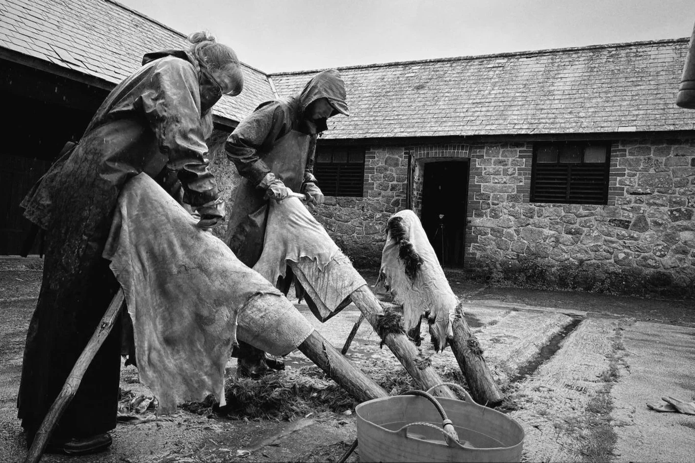 Two people working hides in a courtyard, with a brick building in the background. It is raining.