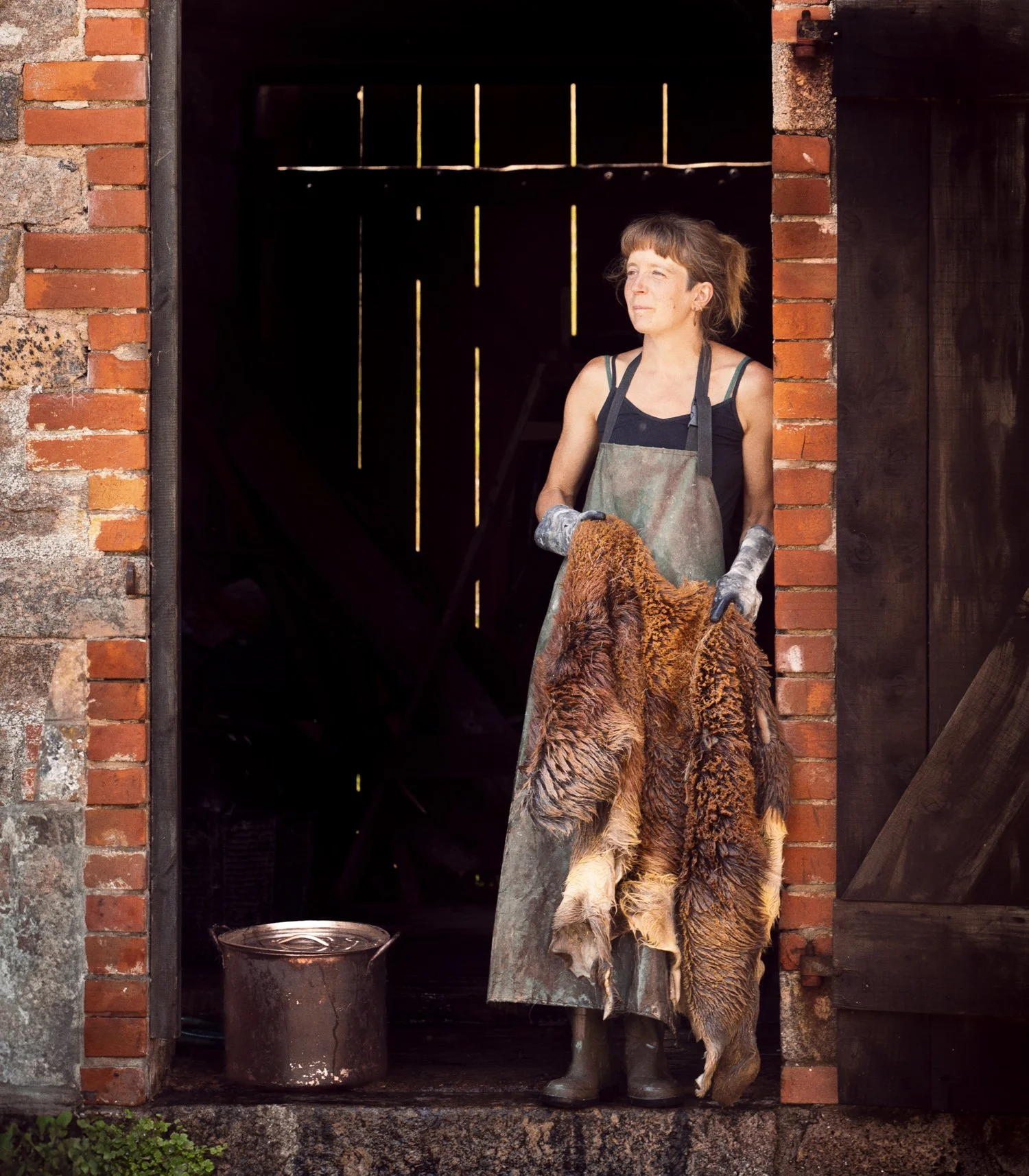 A woman standing in a barn doorway, holding a large animal hide. She is wearing a dark sleeveless top, an apron, and gloves. There is a metal bucket on the ground beside her.