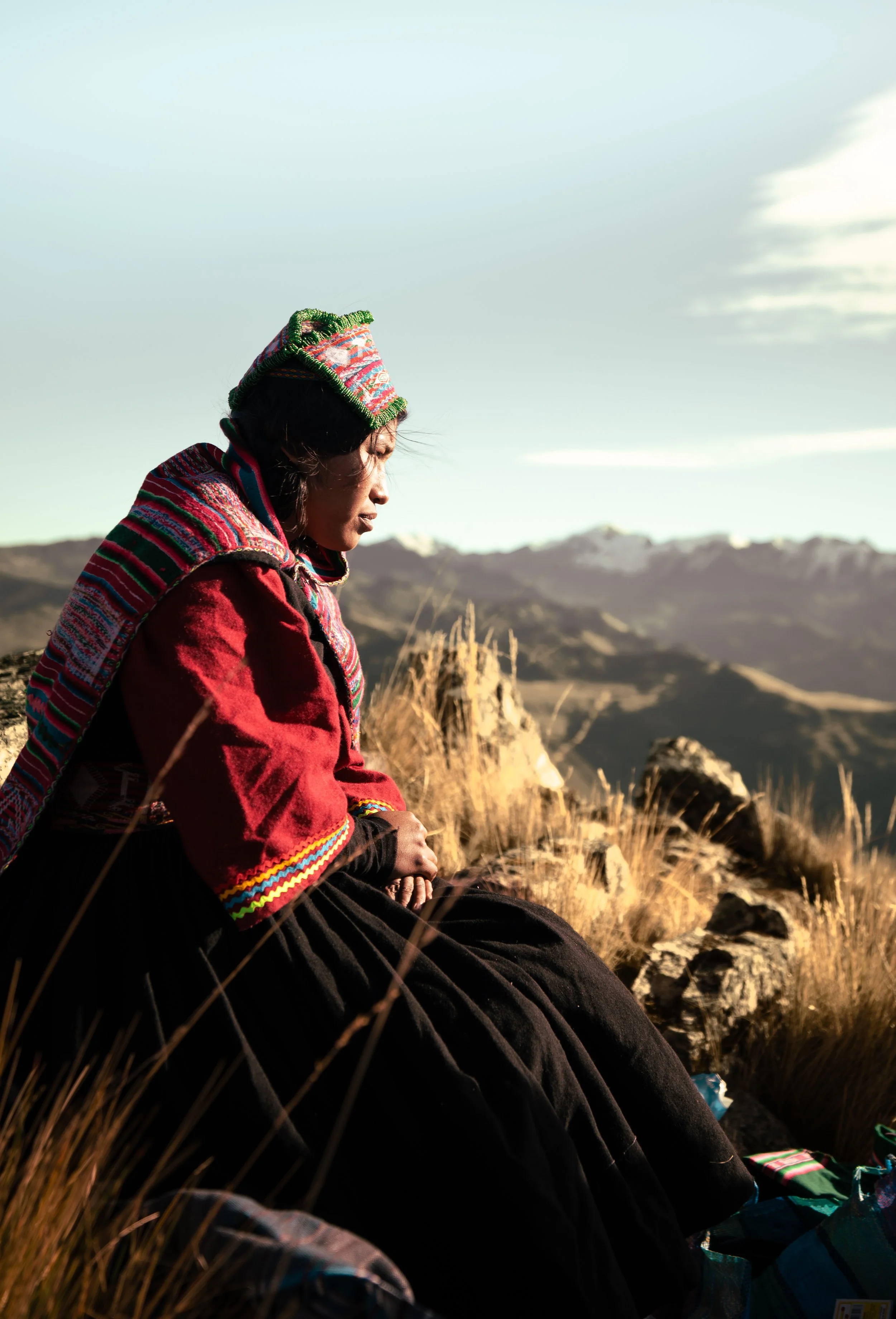 A woman wearing traditional Andean clothing sitting outdoors on a rocky hillside, with mountains and a partly cloudy sky in the background.