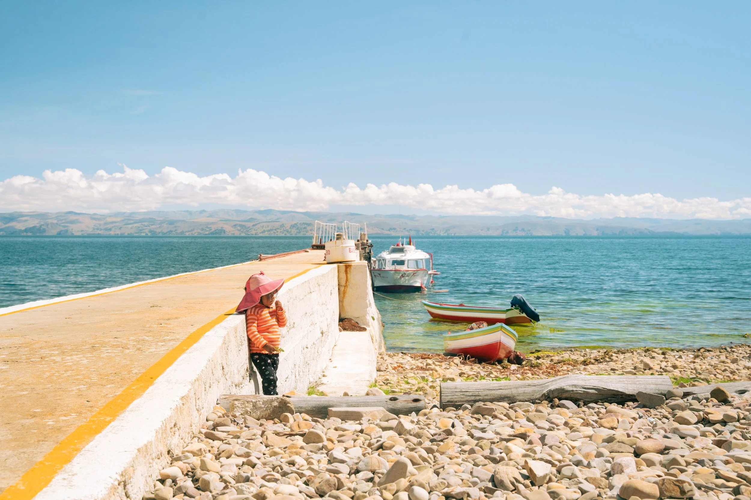 A young girl wearing a pink sunhat and striped clothing leans against a white dock wall by a lake, with multiple boats floating on the water, surrounded by rocky shoreline and distant hills under a bright blue sky with some clouds.