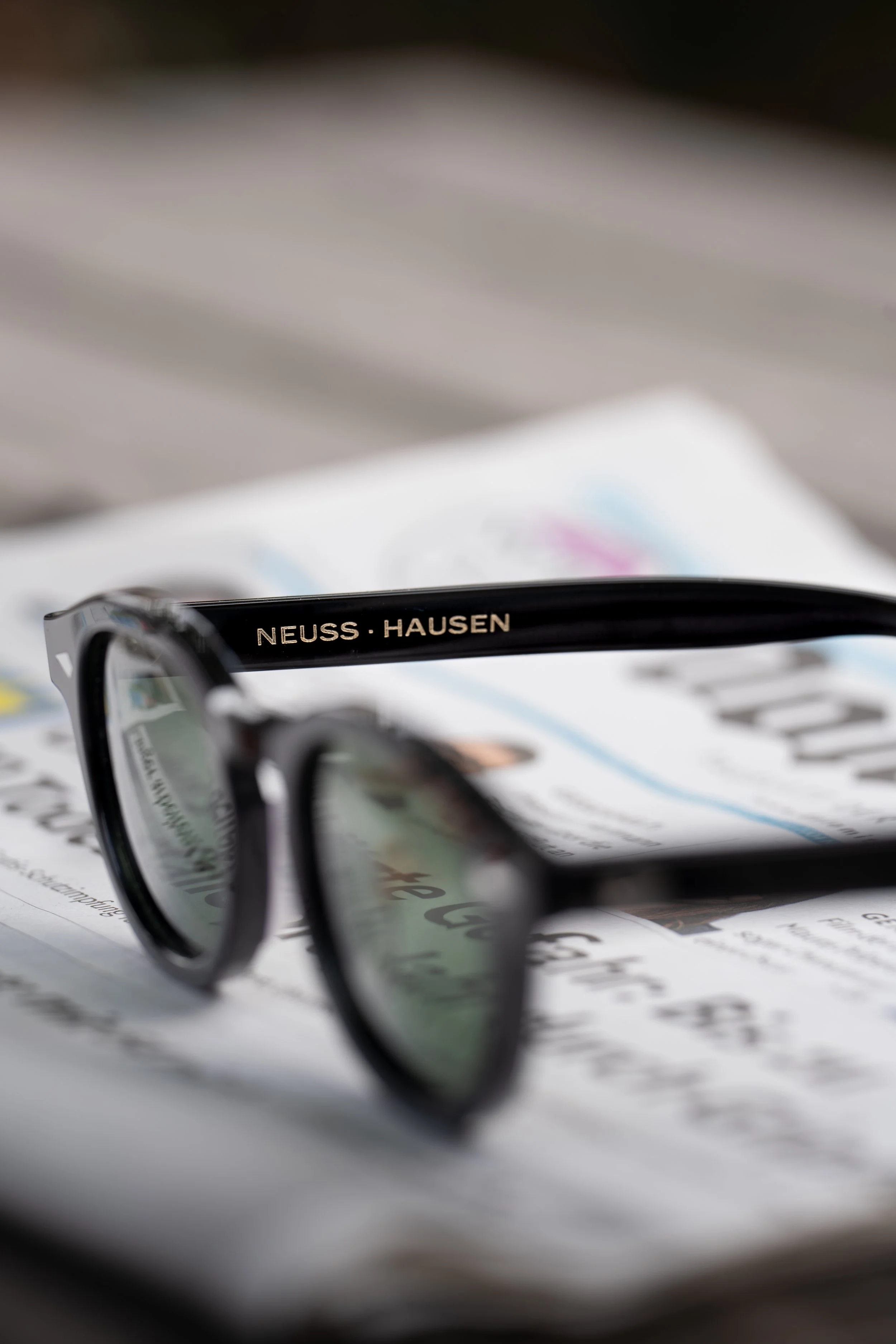 A pair of black eyeglasses with the text 'NEUSS·HAUSEN' on the arm, resting on a printed paper or document.