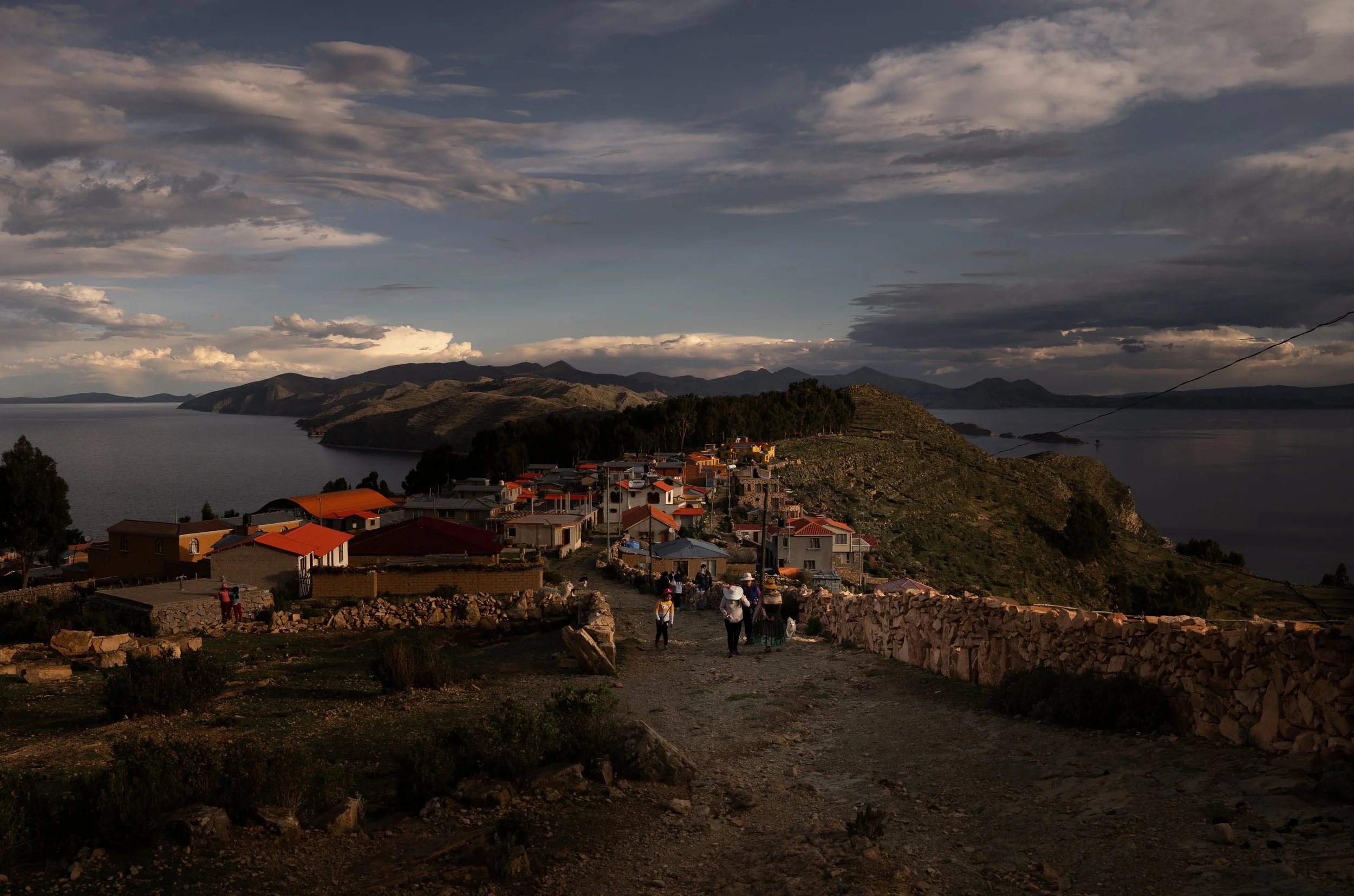 Hilly lakeside village with a few houses, a group of people walking along a dirt path, and mountains in the distance under a cloudy sky.