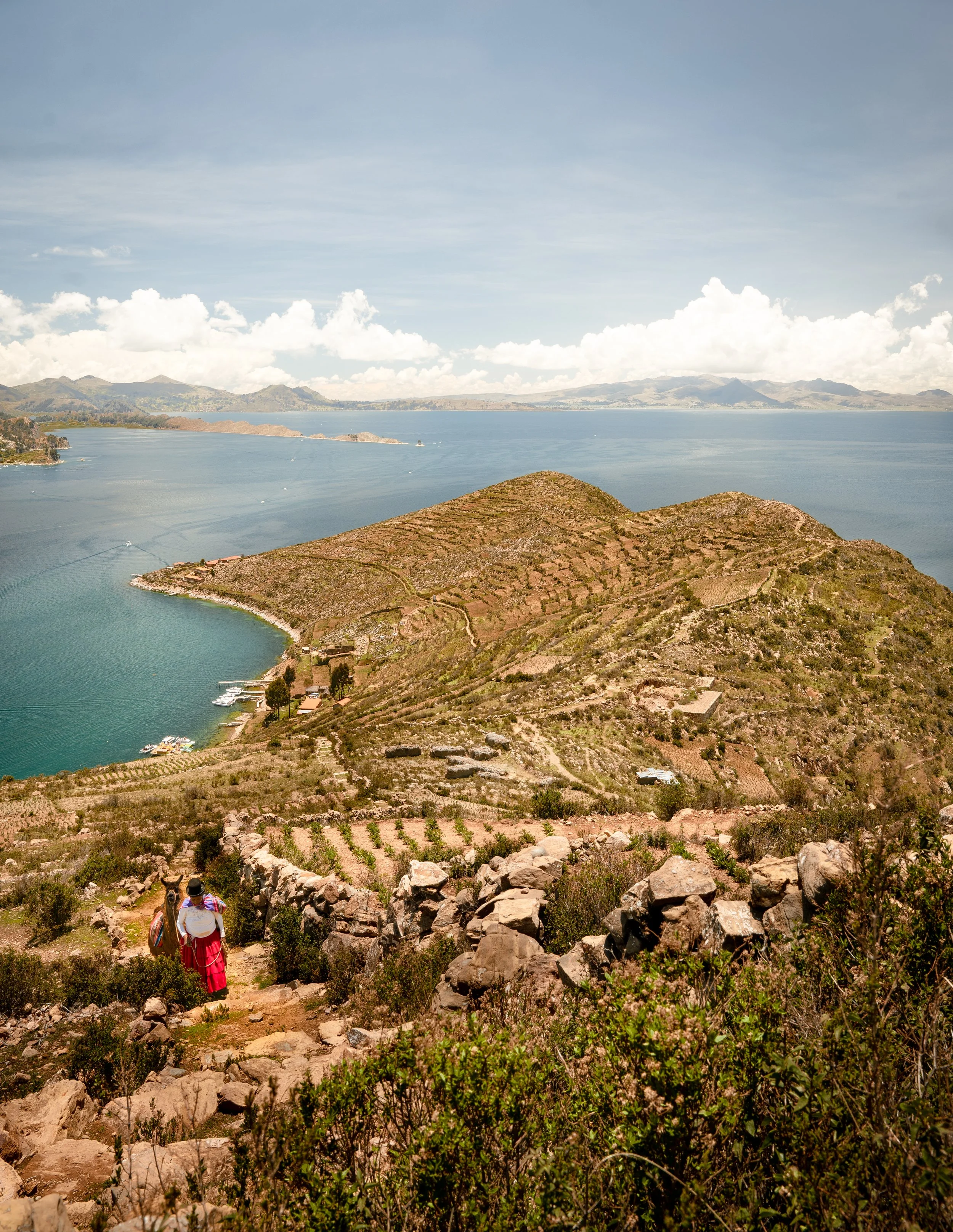 Two women with backpacks walking along a rocky trail through hills with vineyards, overlooking a large body of water and distant mountains under a partly cloudy sky.