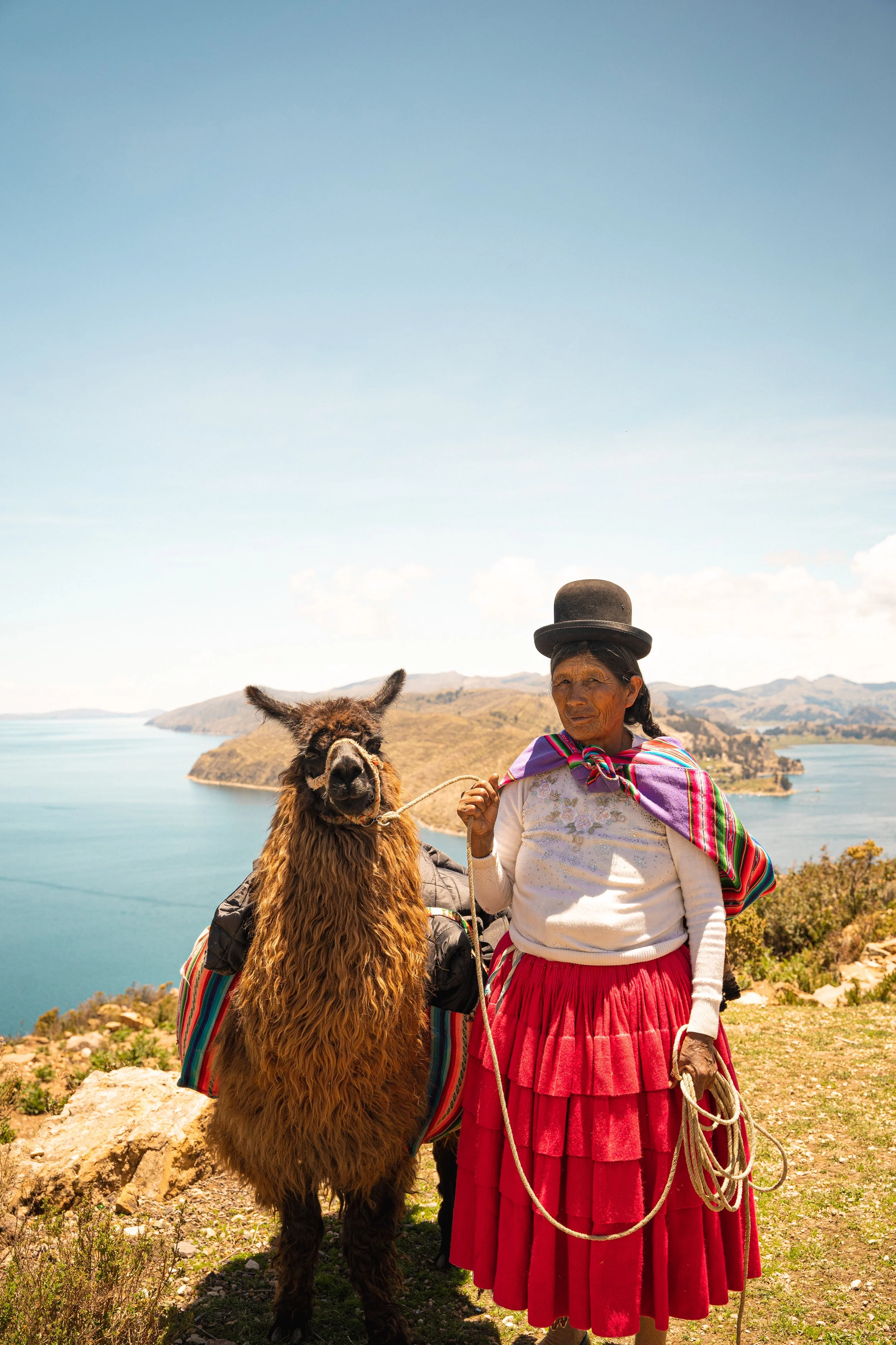 A woman dressed in traditional Andean clothing standing outdoors near a llama, with a lake and mountains in the background.