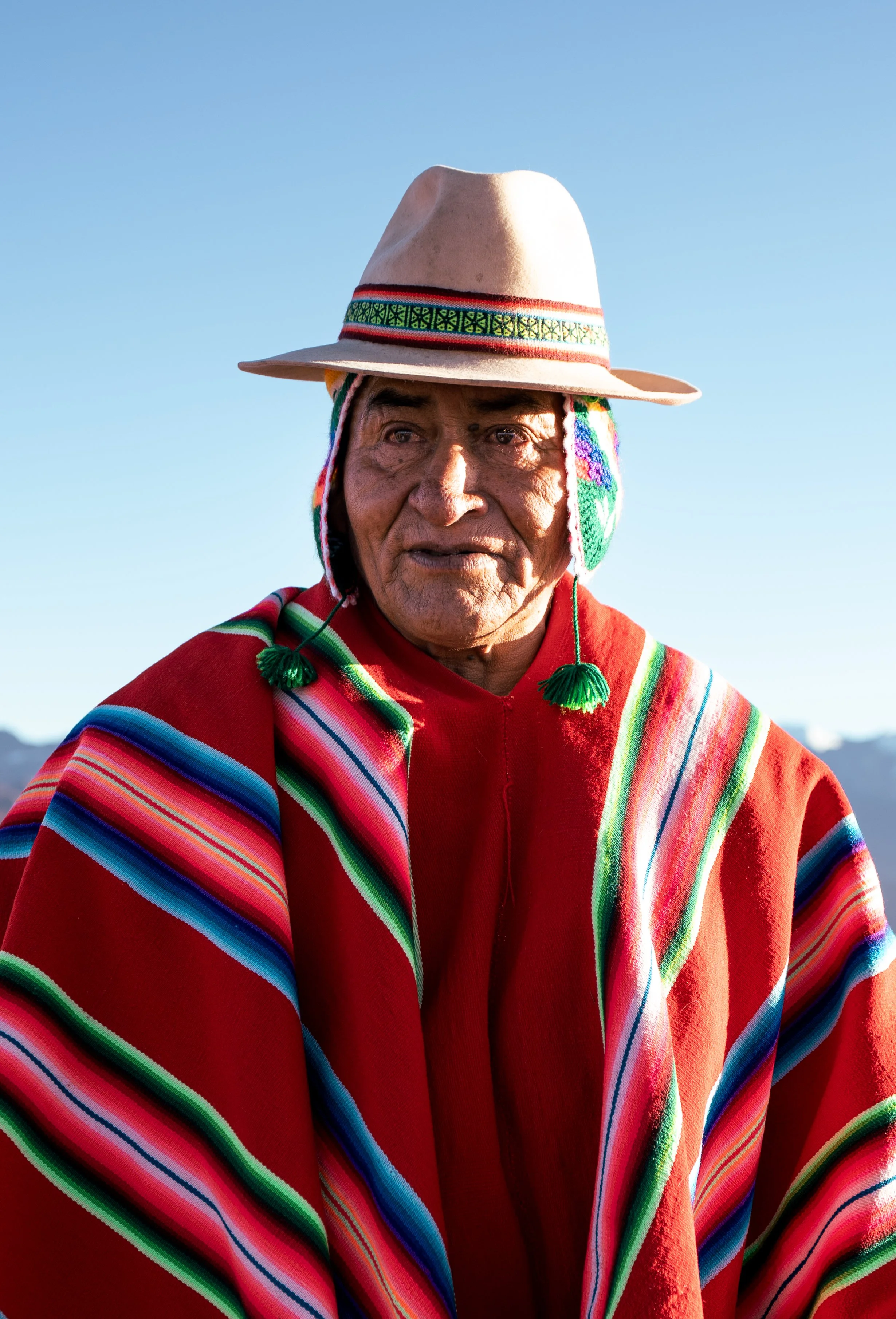 Elderly indigenous man wearing traditional colorful striped shawl, a beige hat with a decorative band, and ear ornaments, standing outdoors with a clear blue sky background.