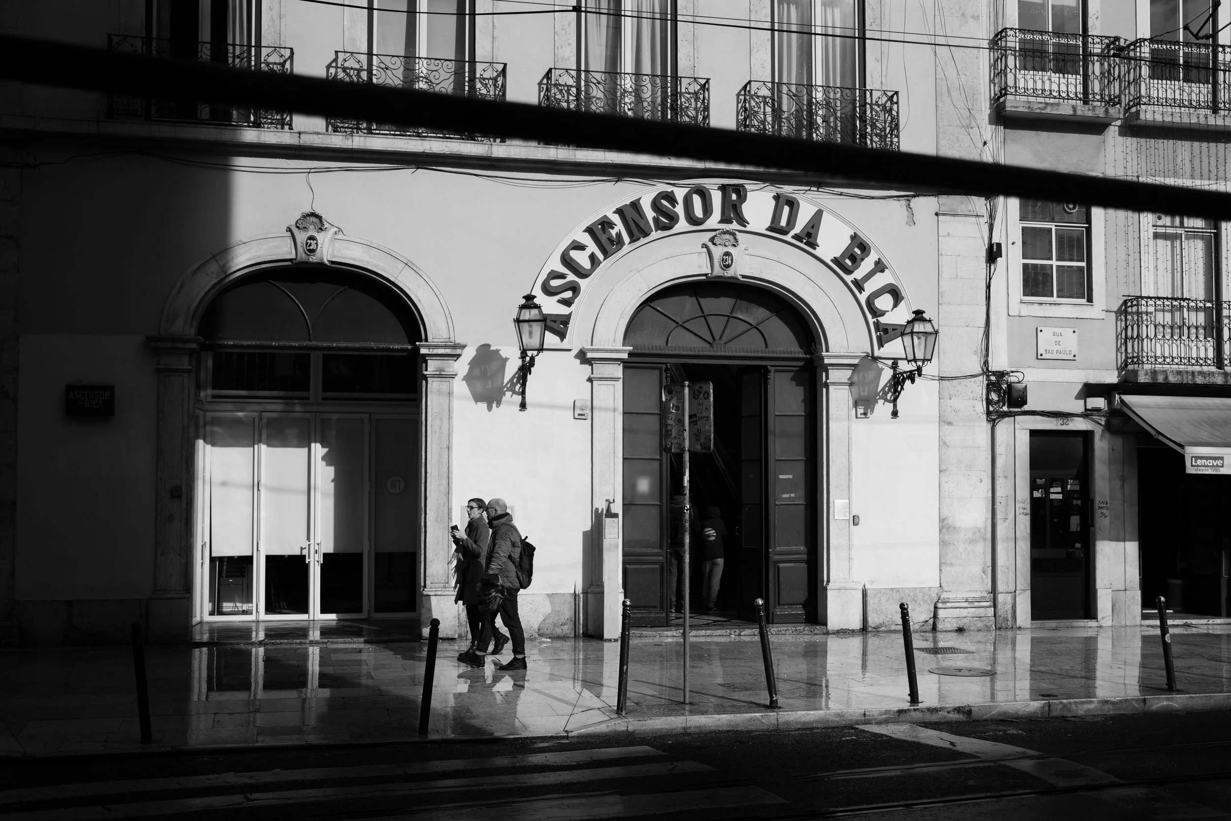 Black and white photo of the facade of a building with a sign that reads 'Ascensor da Bloca'. Two people are walking on the sidewalk in front of the building, with puddles on the ground reflecting their image.