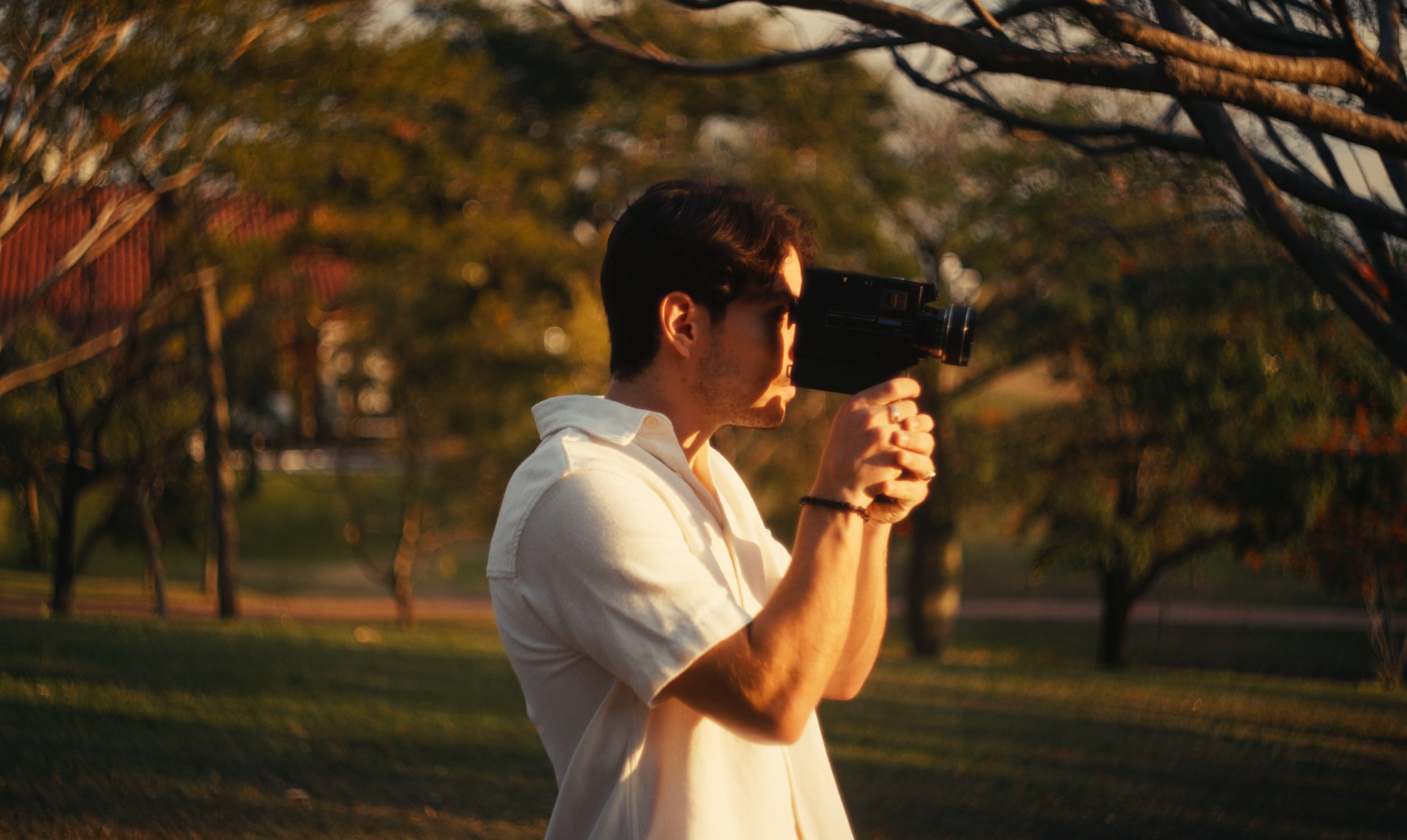 A young man in a white shirt holding a vintage video camera, aiming it towards the right, outdoors during sunset with trees in the background.