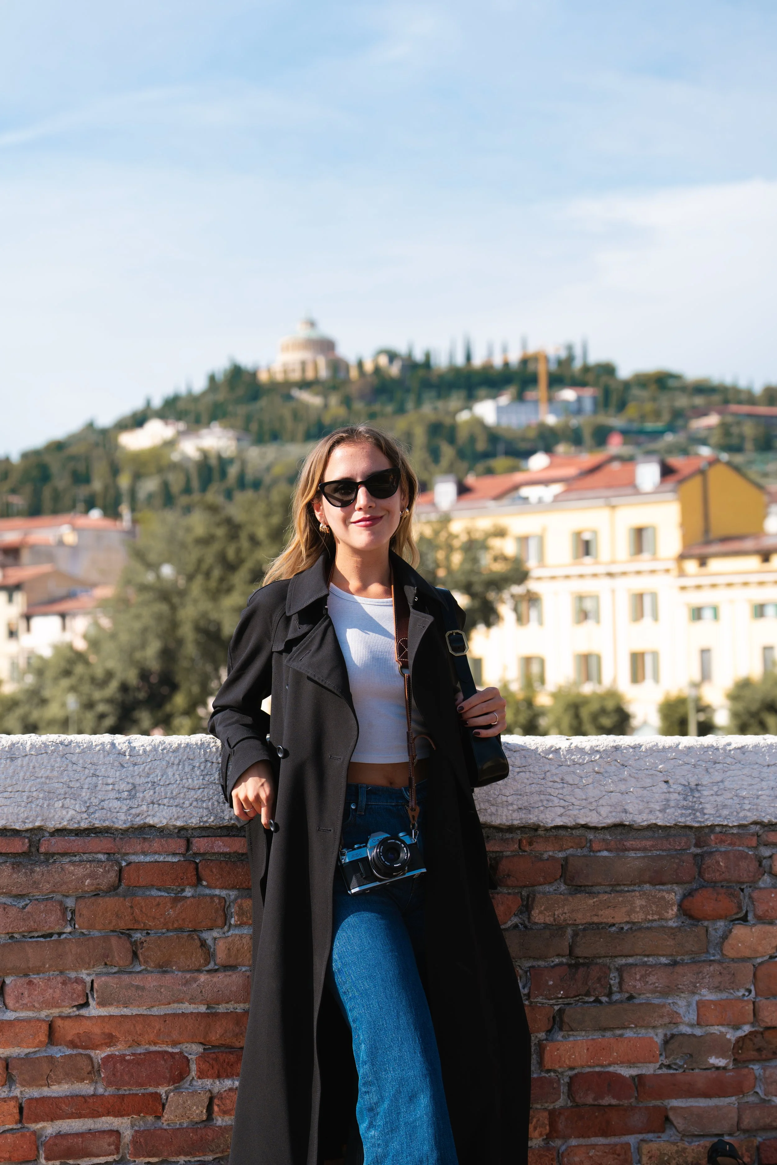 A woman with long blonde hair wearing sunglasses, a black coat, and a white top, standing outdoors in front of a brick wall with a city background including buildings, trees, and a hilltop structure under a clear blue sky.