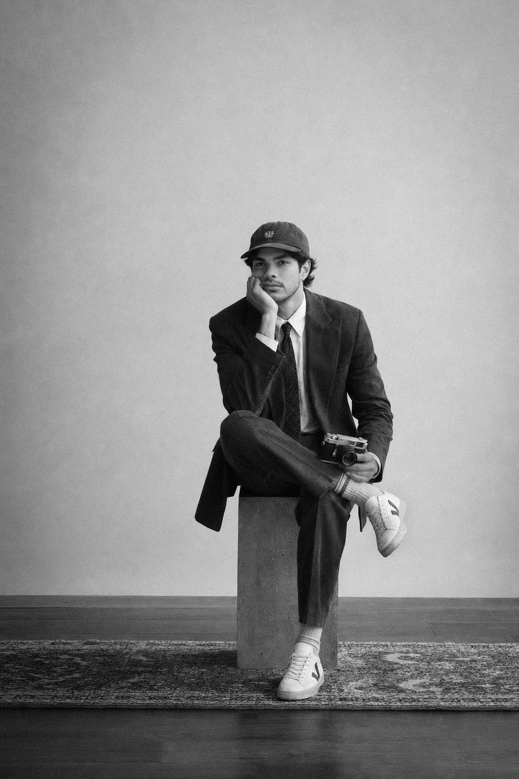 Black and white photo of a young man sitting on a small block, wearing a suit and sneakers, with a camera in his right hand, and resting his chin on his left hand, looking contemplative.