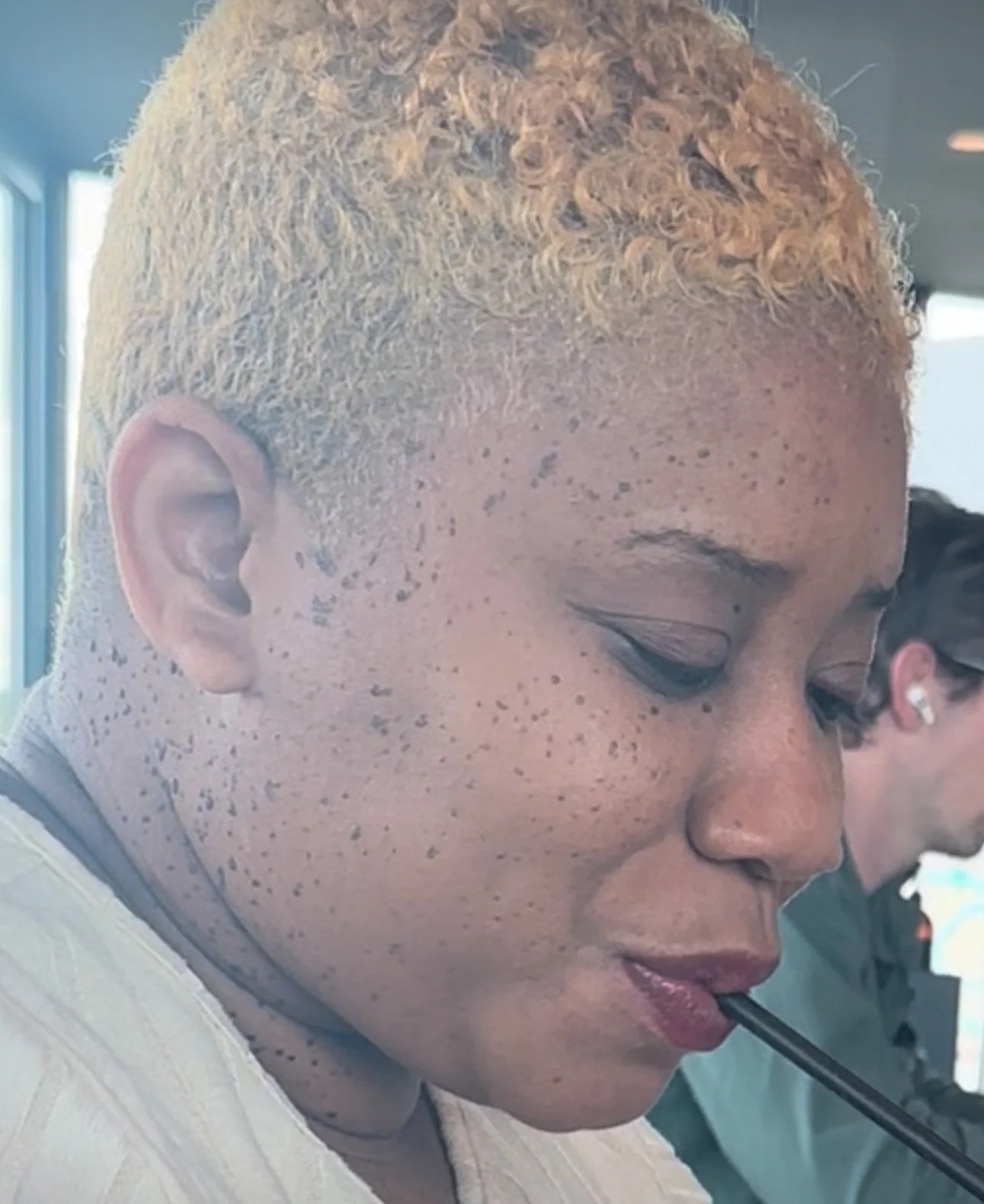 Profile of a woman with short, curly, blonde hair, freckles, and light makeup, sitting indoors with her eyes closed, holding a black straw in her mouth.