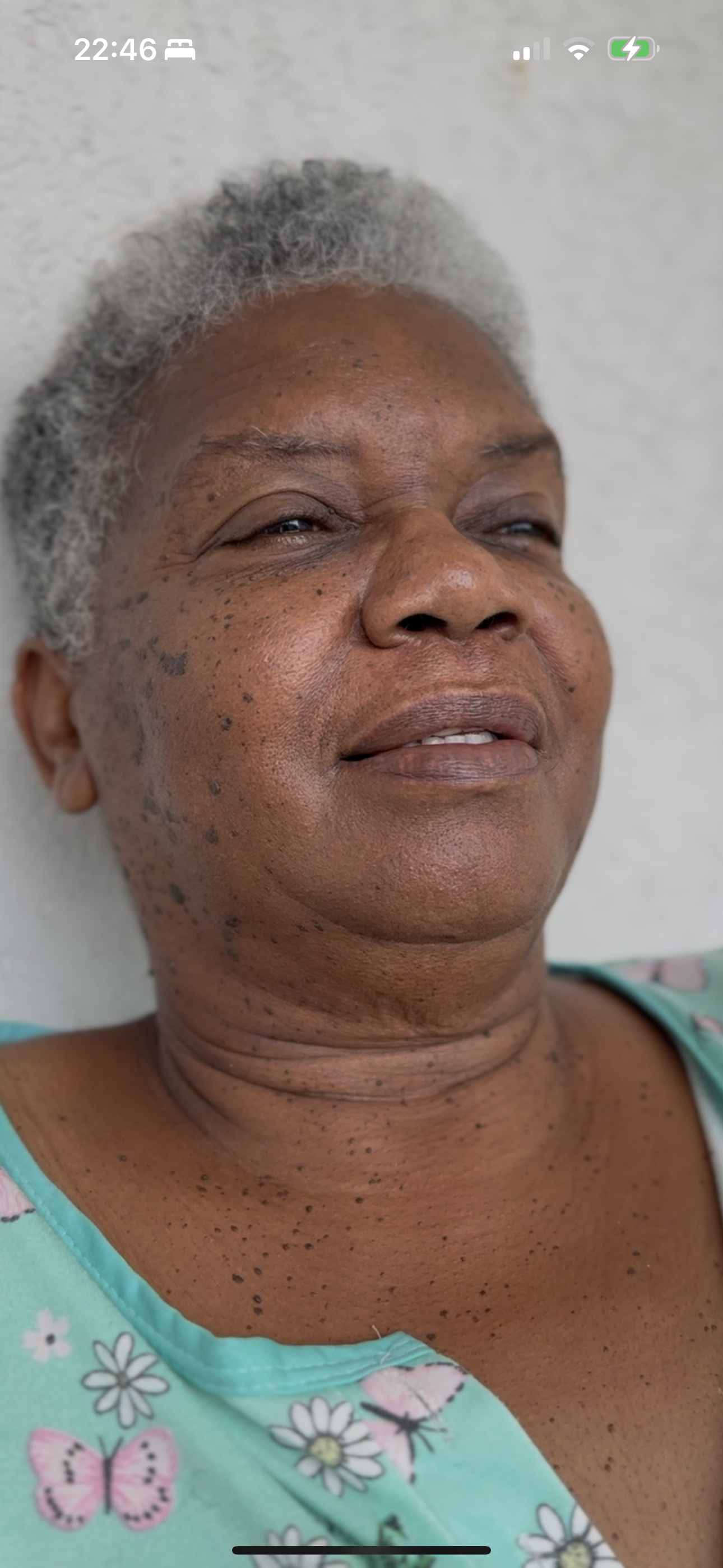 Close-up of an elderly woman with gray hair, smiling gently, wearing a turquoise top with butterfly and flower patterns, positioned against a plain white background.