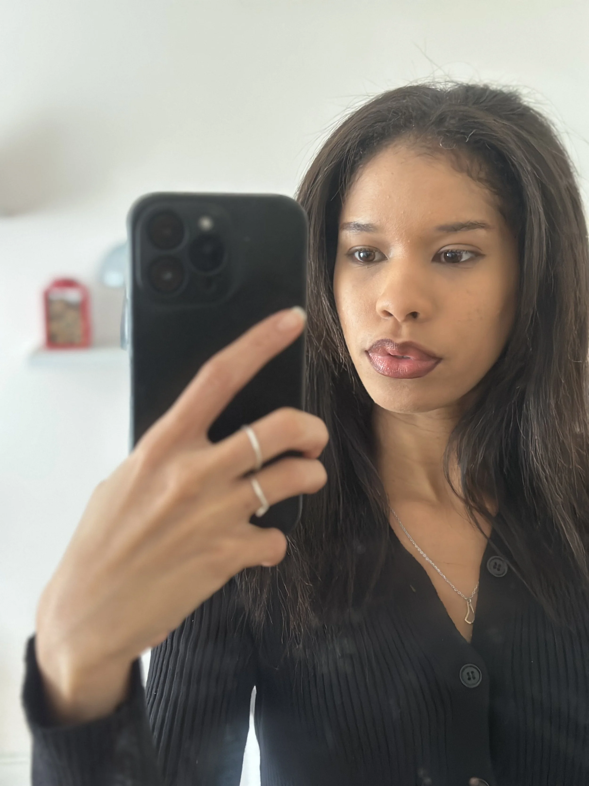 Woman taking a selfie in a mirror, wearing a black button-up shirt, with long dark hair and makeup visible.
