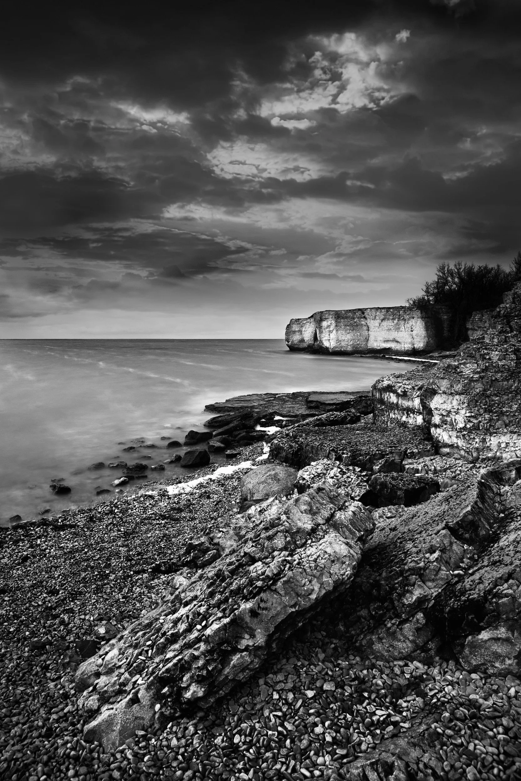 Black and white photo of a rocky cliffside by a lake or sea, with a cloudy sky overhead and trees on top of the cliff.
