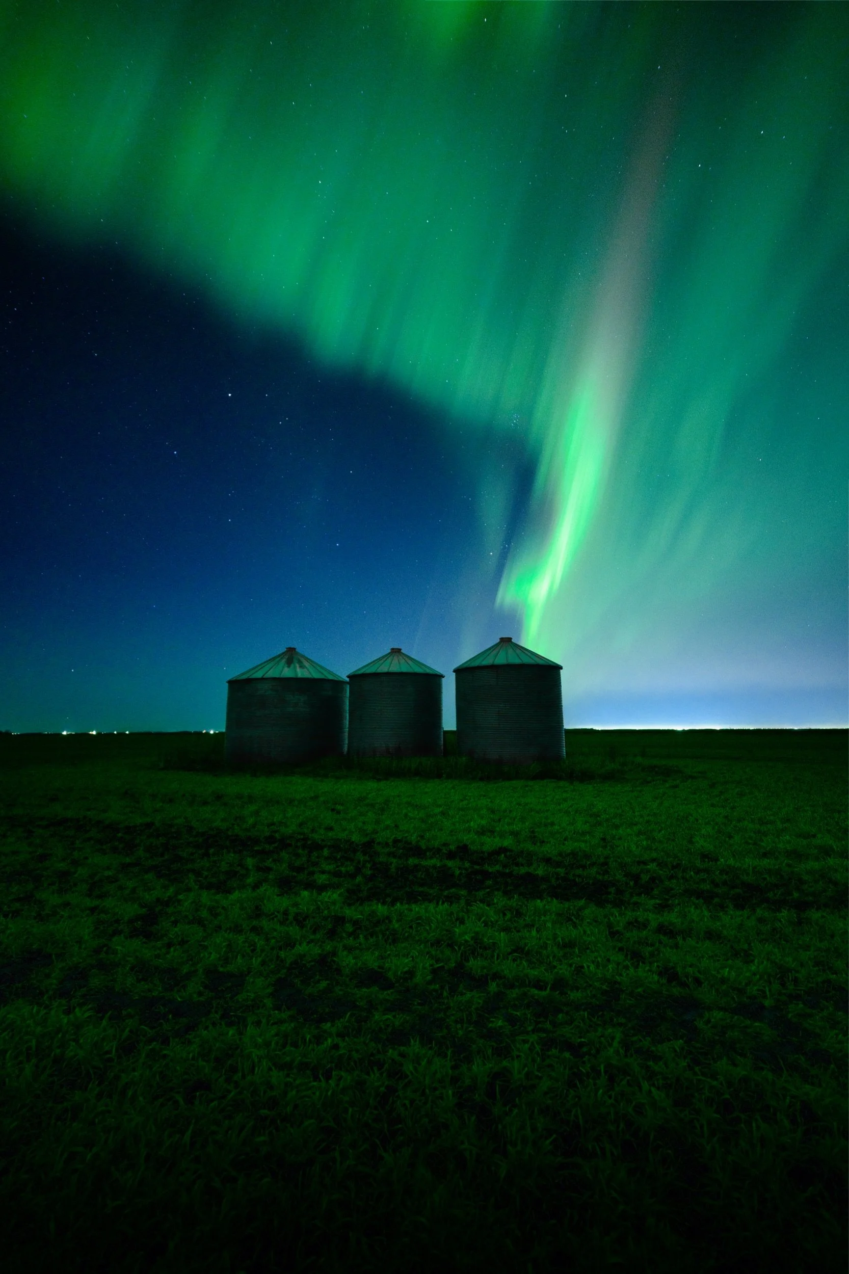 Three grain silos on a grassy field under a night sky with green Northern Lights and stars.