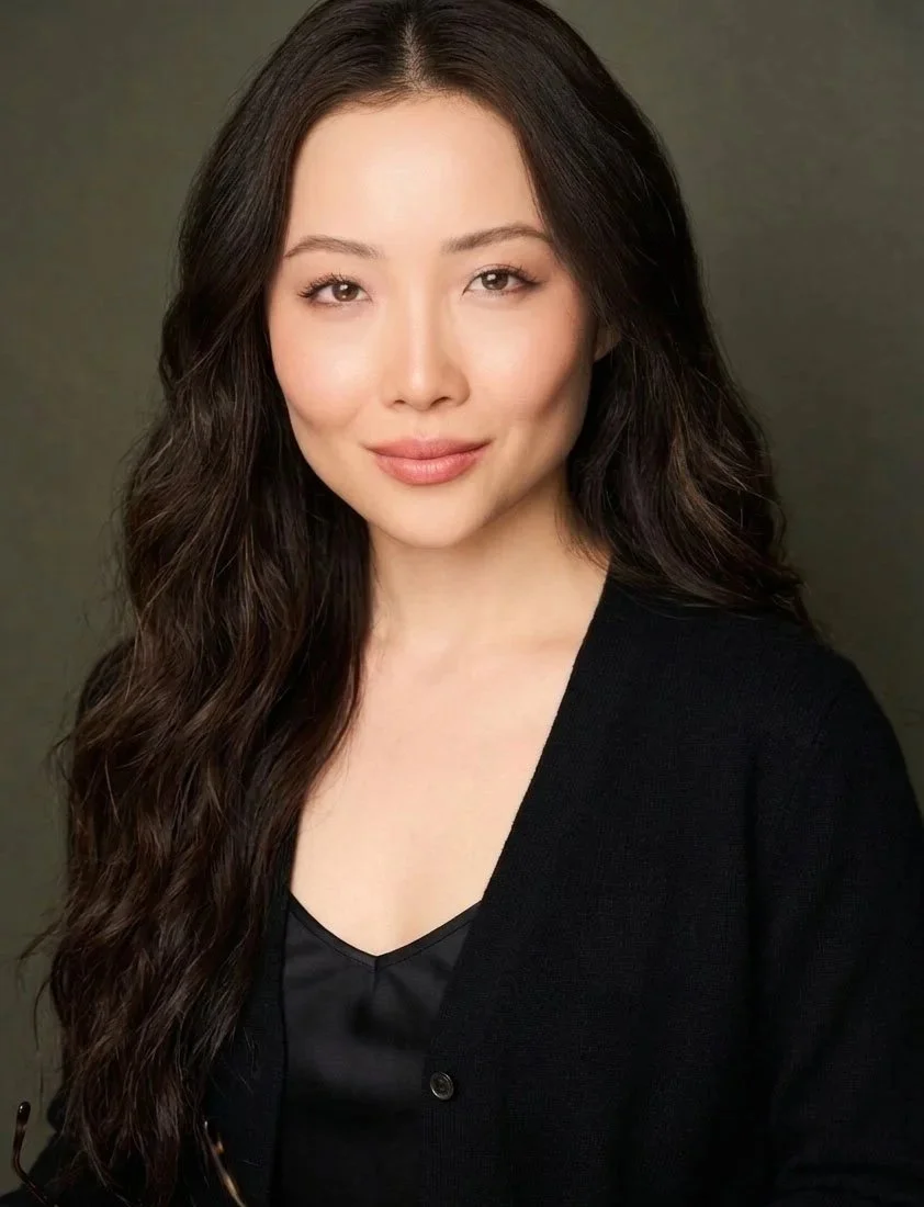 Portrait of a woman with long wavy dark hair, wearing a black top and blazer, smiling subtly against a plain dark background.