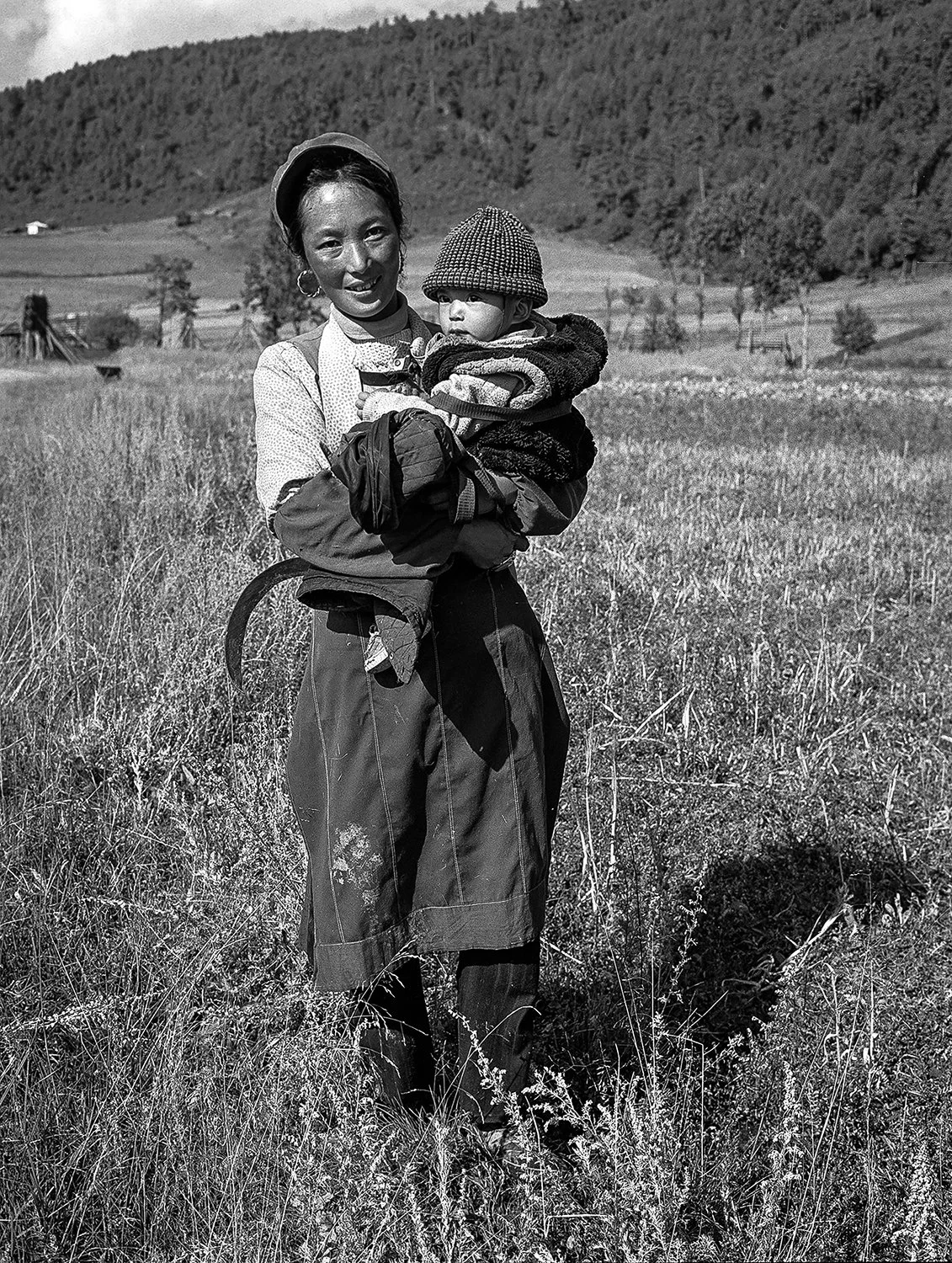 A woman holding a child in a field with mountains and trees in the background, in black-and-white.