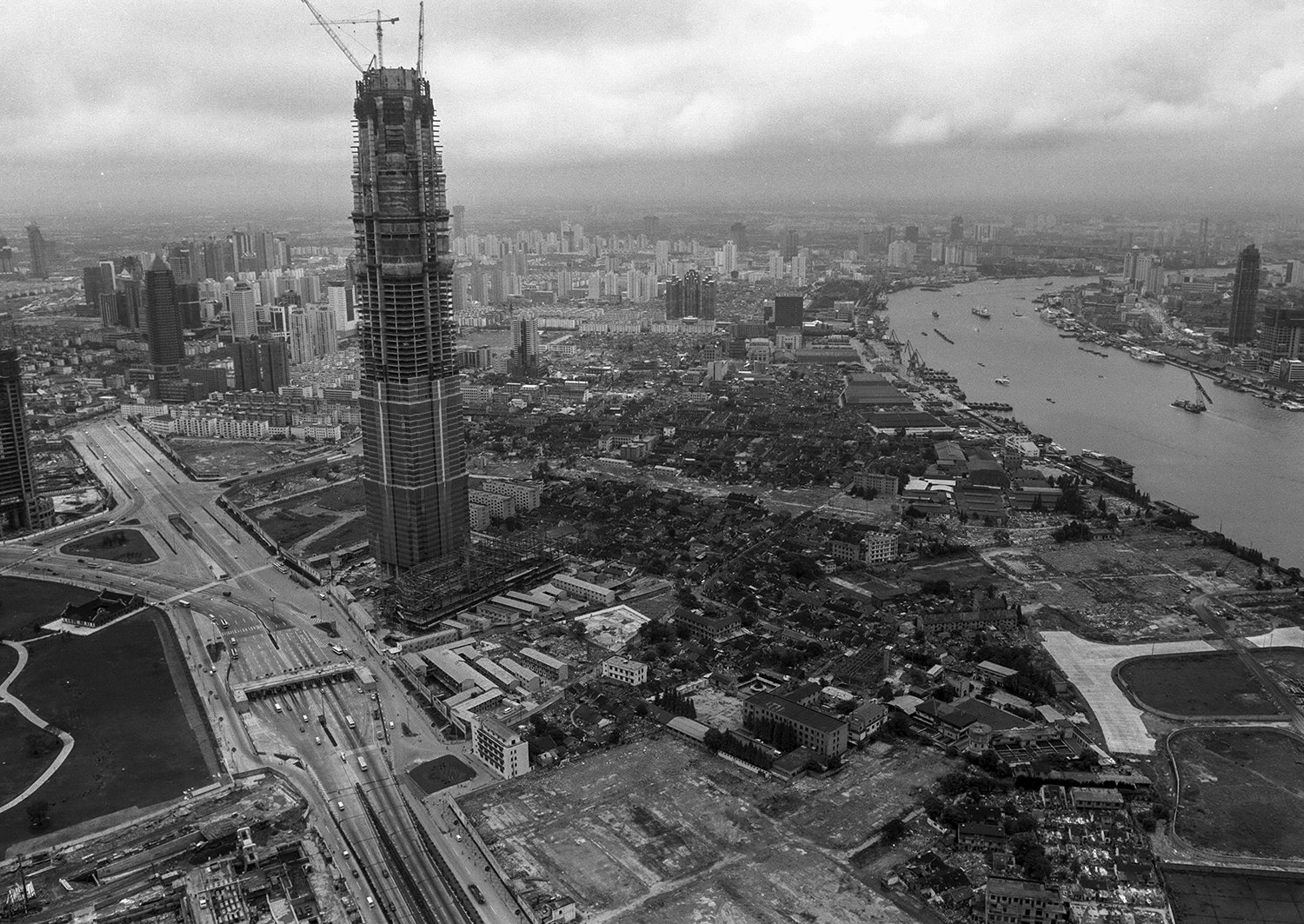 Aerial view of a cityscape with tall buildings, a river, and busy roads. A skyscraper under construction is prominent in the foreground.